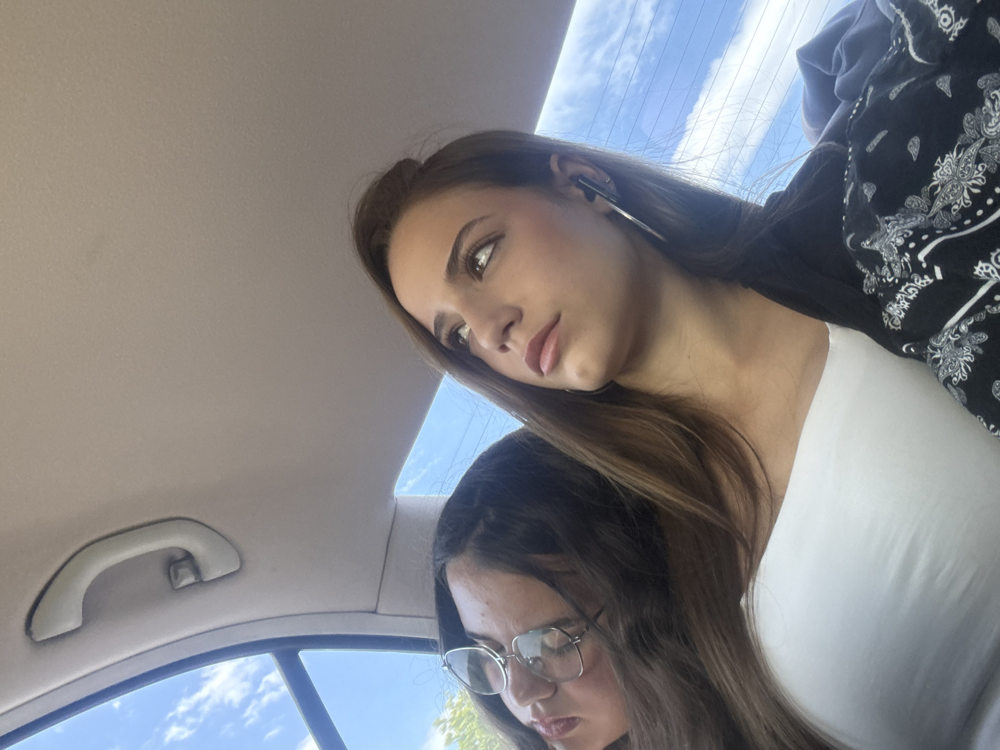The image depicts two young women inside a car, potentially in motion, based on the background view of a partly cloudy sky through the car window. The woman in the foreground is fair-skinned with long brown hair, wearing a white top and a jacket with a black and white paisley print. She has large hoop earrings and appears to be looking downward and to the left. The second woman, positioned slightly behind and below, also has long brown hair and wears glasses. A car grab handle is visible in the upper left corner of the image. The location is not discernible as Gorni Bogrov, Bulgaria.