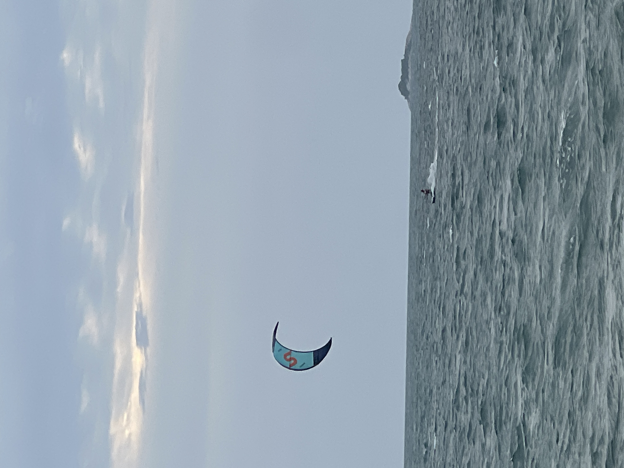 A kite surfer is pictured on the ocean on a cloudy day. The kite is a crescent shape and colored turquoise with a red "S" on it. The kite surfer is a small figure in the distance, wearing red and white. The water is choppy and dark gray. In the far distance on the right, a rocky island is visible. The sky is a pale blue-gray with scattered clouds. The location is Ko Pha-ngan District, Thailand.