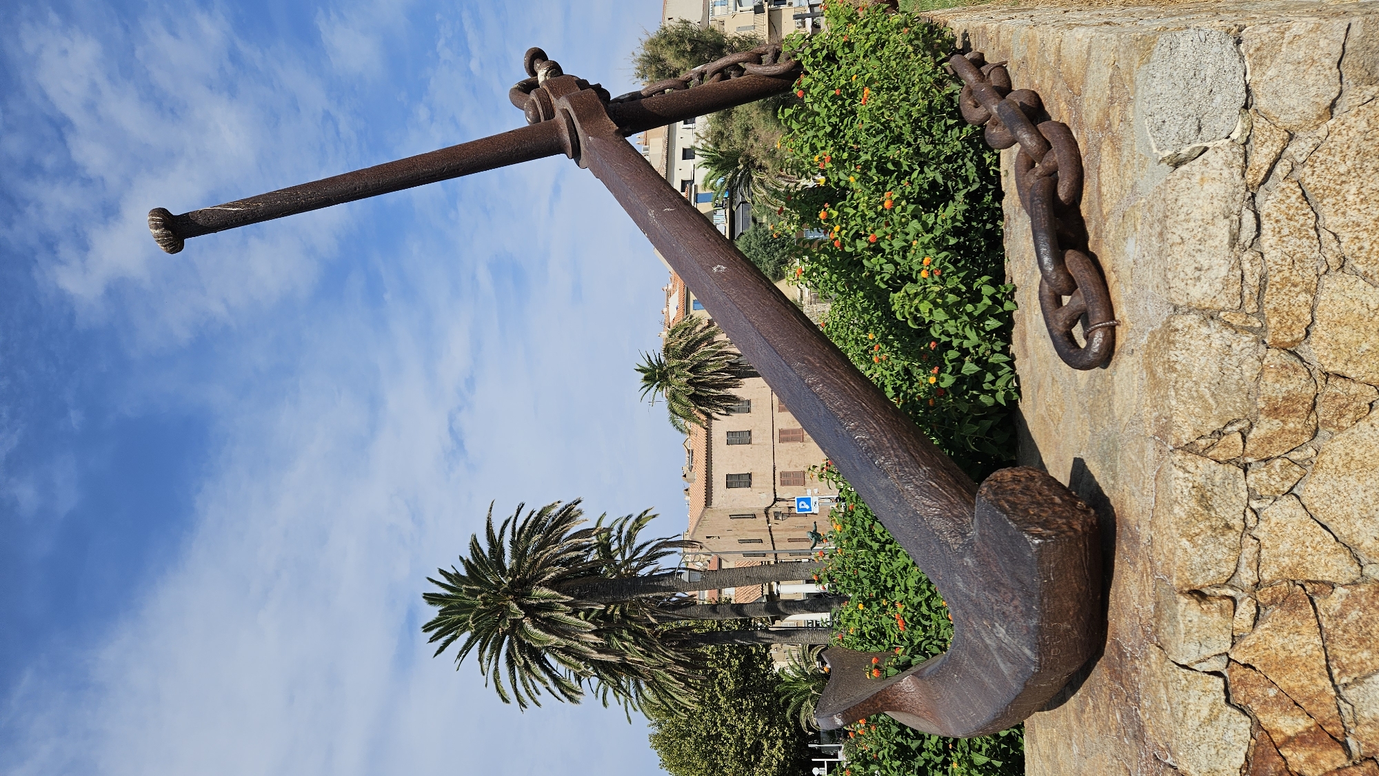 An anchor monument and a detached anchor chain rest on a stone base. The anchor is made of rusted metal and is positioned diagonally with its fluke on the left and its shank extending towards the upper right. A separate section of chain lies on the ground to the right of the anchor. Behind the anchor, a garden with green foliage and orange and yellow flowers is visible. In the background, there are palm trees and buildings with tiled roofs. The sky above is blue with scattered white clouds. The scene appears to be outdoors, likely in a public space in the city of L'Île-Rousse, France, given the architectural style and vegetation.