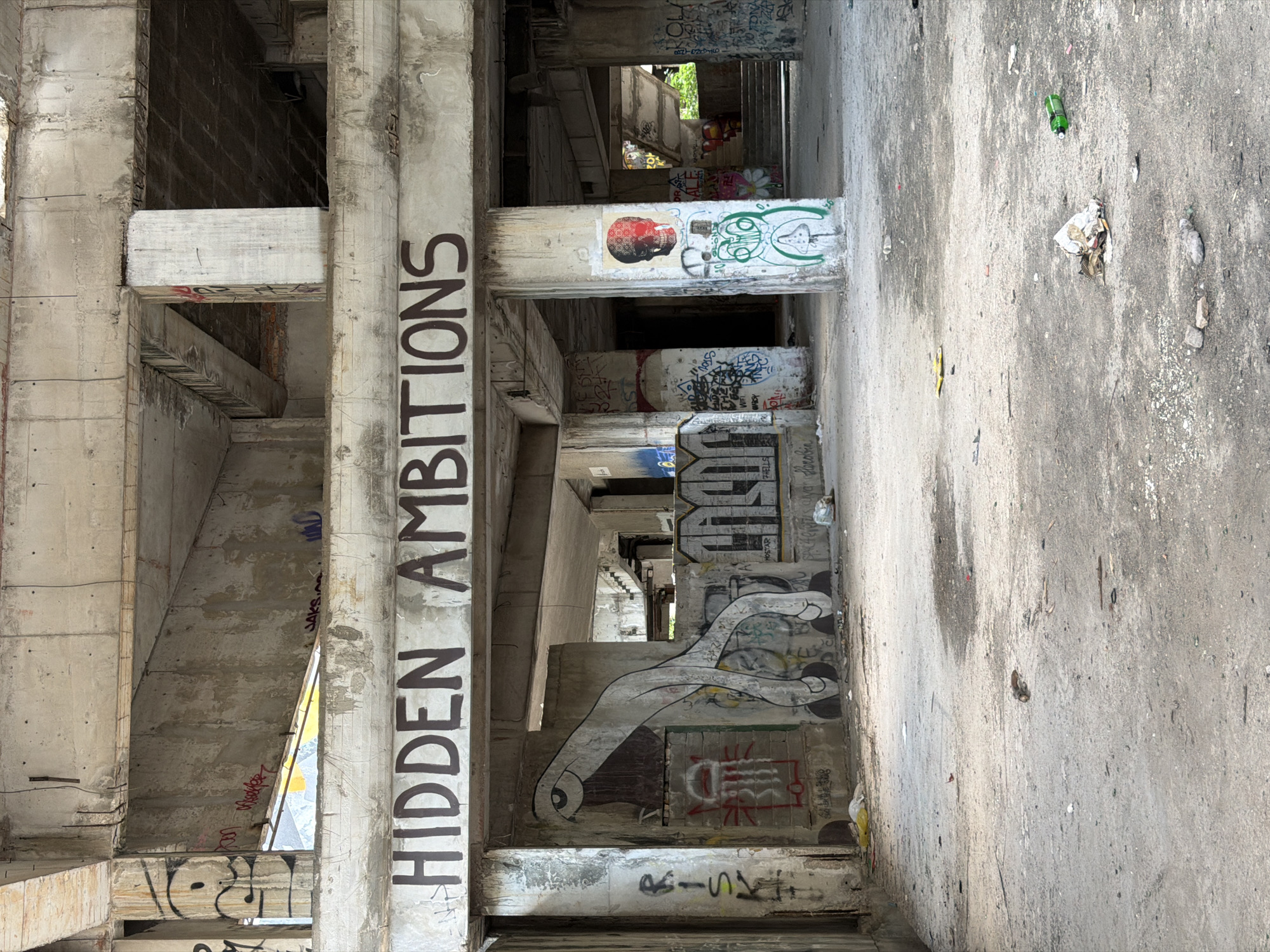 The image shows the interior of an abandoned concrete structure in Mostar, Bosnia and Herzegovina. Large, graffiti-covered concrete beams and columns dominate the scene. A prominent phrase, "HIDDEN AMBITIONS," is spray-painted in dark letters across a horizontal beam. Various graffiti tags and images adorn the walls and pillars, including a stylized eye and a skull with red eyes. The ground is covered in concrete dust and debris, with scattered litter such as plastic bags, paper, and a green glass bottle. The overall impression is one of decay and urban exploration.