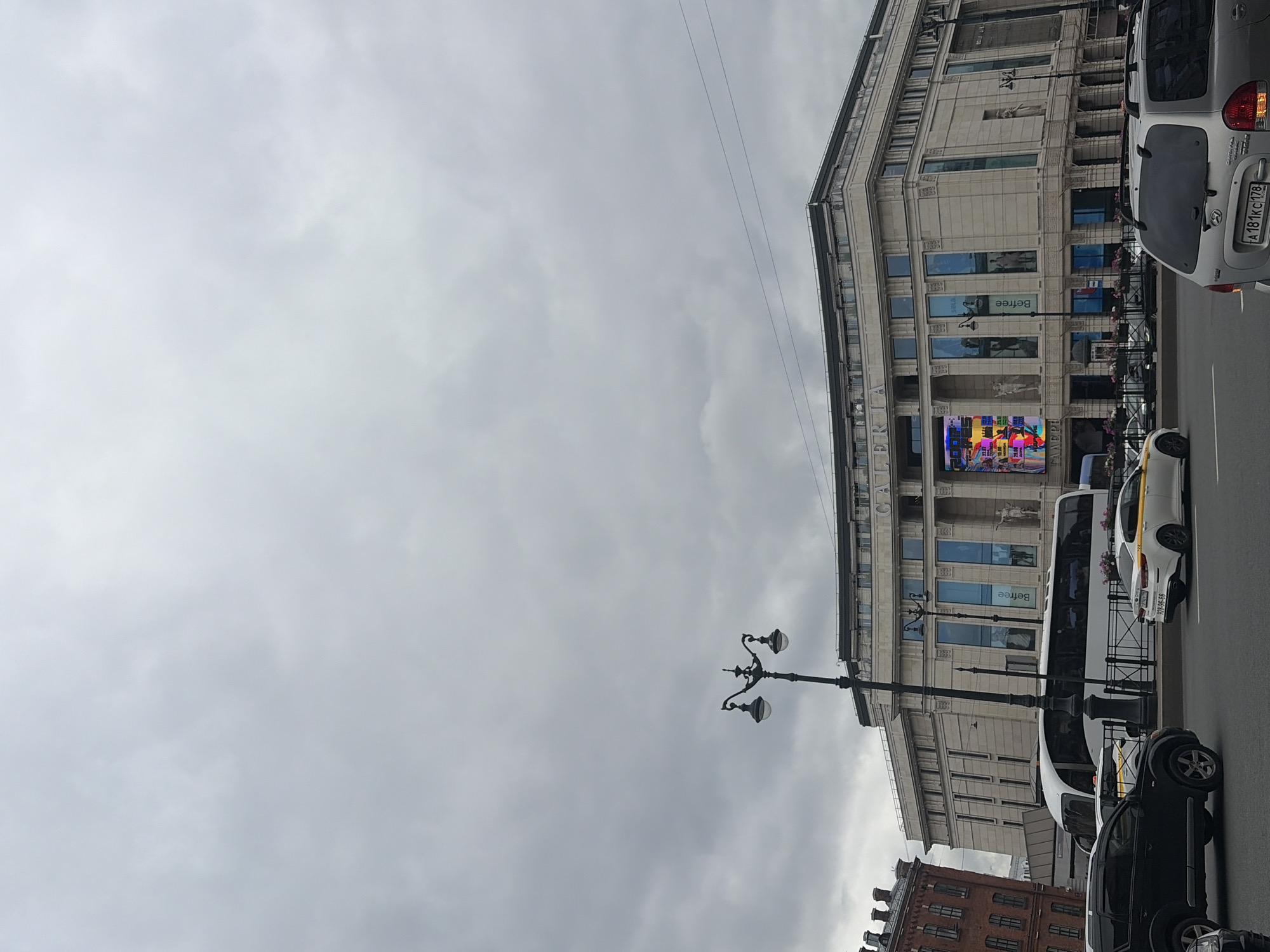 The image shows a street scene in front of the Nevsky Center shopping mall in Saint Petersburg, Russia. The mall, officially known as Galeria, is a large, modern building with a curved facade and a prominent digital billboard. The billboard is displaying a colorful advertisement. In front of the mall, there are several cars, including a white bus, a black SUV, and two yellow and white taxis. People are visible inside the bus. The sky is overcast and gray, suggesting cloudy weather. The scene is set during the day.