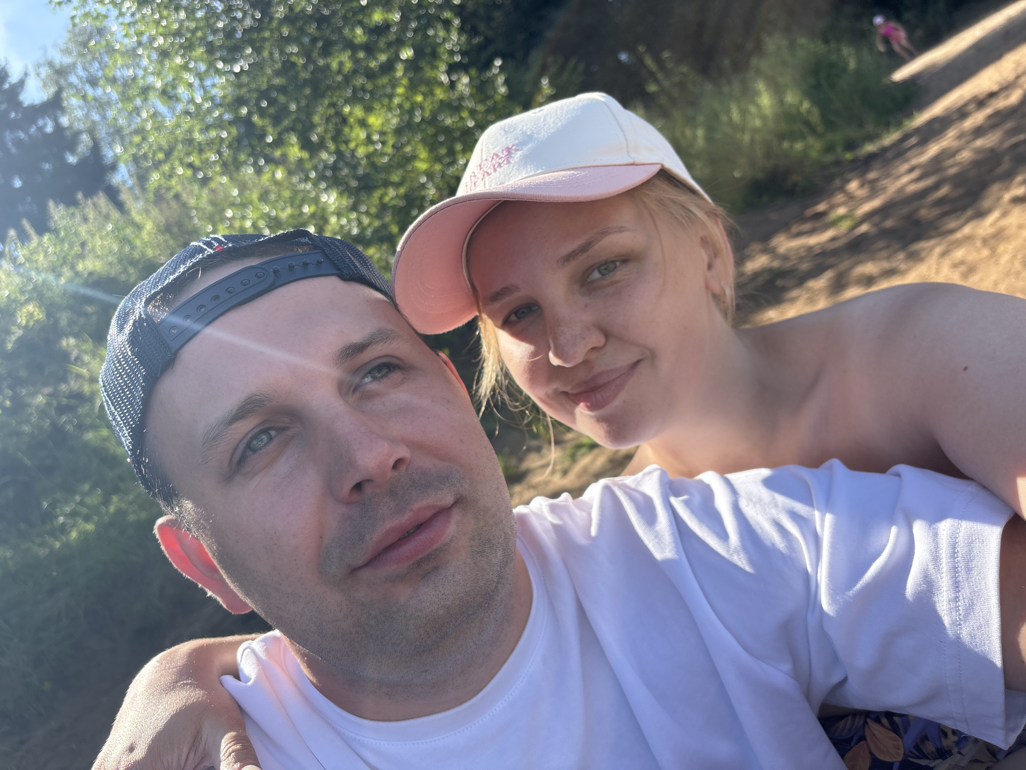 This is a selfie taken outdoors, likely on a sunny day, with a couple. The man is in the foreground on the left, wearing a white t-shirt and a dark baseball cap. He is smiling and looking at the camera. The woman is positioned behind and to the right of the man, also looking towards the camera and smiling. She is wearing a white baseball cap with pink trim and text on it. The background consists of blurred green foliage and dappled sunlight, suggesting a natural setting such as a park or a wooded area. The overall impression is one of a relaxed and happy moment shared between the two individuals. There is no discernible indication of the city of Yaroslavl, Russia, or any specific urban elements within the image.