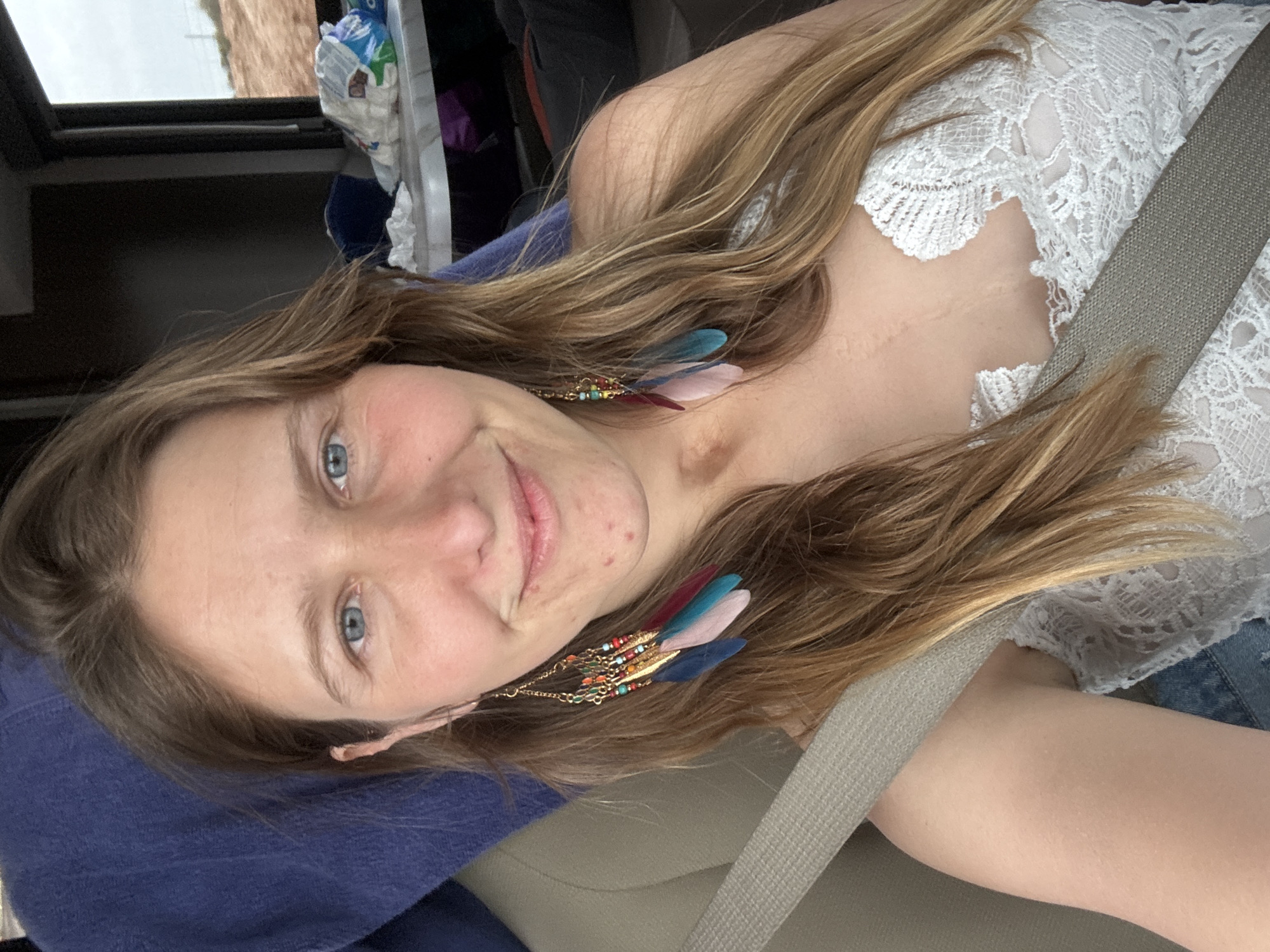 A woman is pictured inside a car. She is wearing a white lace top and has a seatbelt across her torso. The woman has long, wavy hair with a light brown hue, and she's wearing dangling earrings with feathers. Her face shows fair skin and blue eyes, with a slight smile. The car interior includes a dark blue cushion or fabric on the left, and the back of a seat can be seen in the background, along with various items. The location is not discernible based on the image.