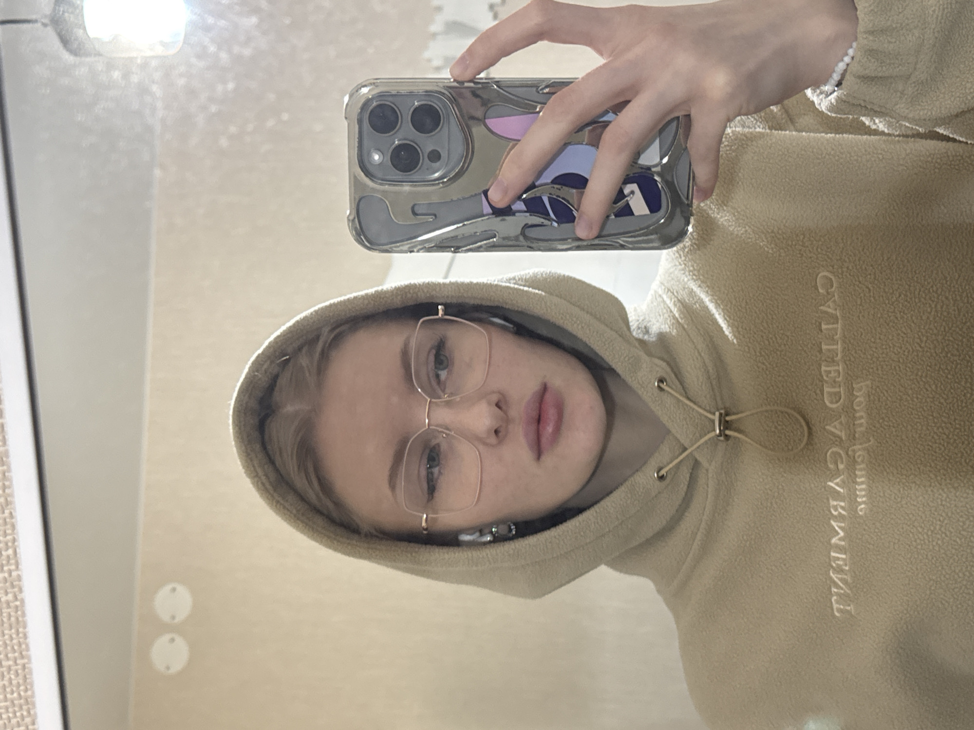 A young woman takes a selfie in a mirror. She is wearing glasses and a light beige hoodie with the name "Boy Tomorrow" written in reverse, likely due to the mirror reflection. She is holding a smartphone with a patterned case. The background appears to be a wall, possibly in a room. There are no explicit indications of the city of Perm, Russia.