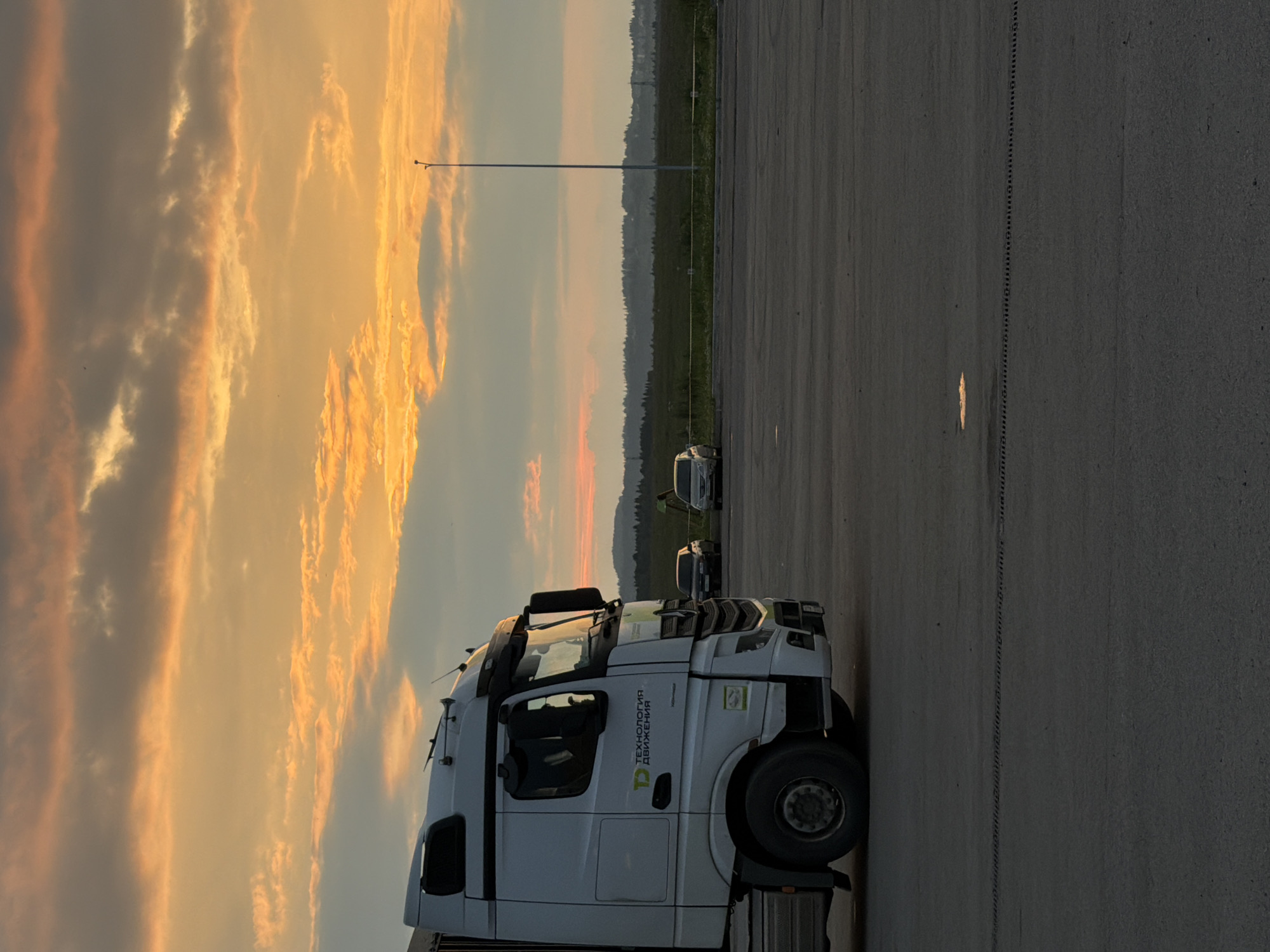 A white semi-truck is parked on an asphalt lot. The truck is in the foreground, taking up the left side of the frame. The truck has "ТЕХНОЛОГИЯ ДВИЖЕНИЯ" written on its side in green and white lettering. The truck is facing towards the right of the frame. In the background, there is a line of trees and hills. The sky is a dramatic mix of orange, yellow, and grey clouds, indicating sunset or sunrise. A single lamppost stands in the background, to the left of the truck. Two smaller vehicles, possibly cars, are parked further back in the lot, to the right of the truck. The asphalt lot appears to be empty except for the parked vehicles. The lighting suggests it is either early morning or late evening. The setting appears to be an outdoor lot, possibly a rest stop or a parking area for vehicles. The overall mood is calm and serene, highlighted by the beautiful sky.