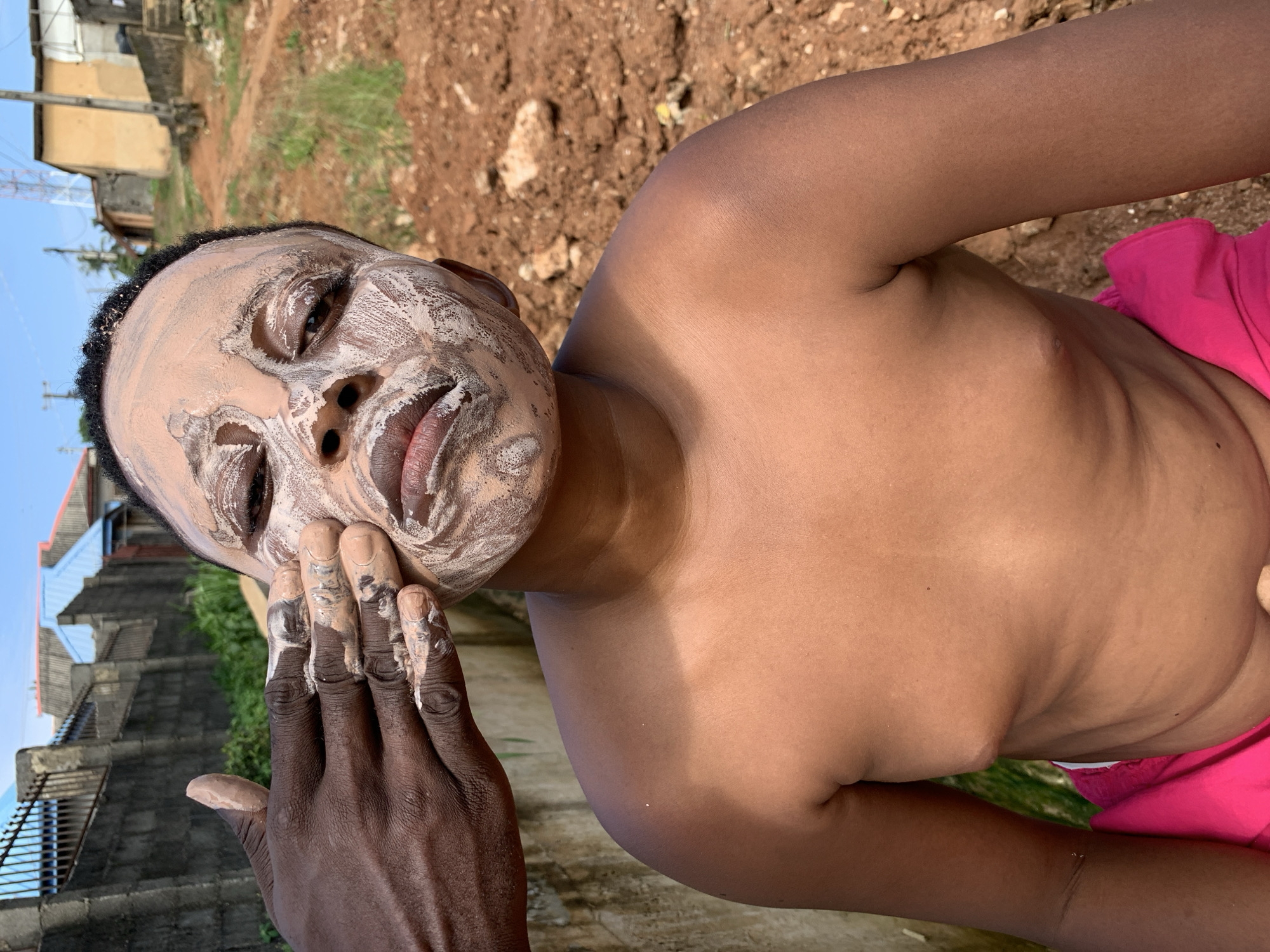 This media file shows a close-up of a child's face being covered in a light-colored substance, possibly clay or makeup, with a hand smearing it on. The child has dark skin and is looking towards the camera with a neutral expression. The background is out of focus but appears to be an outdoor setting with brown soil and some green vegetation. In the background, there are hints of buildings, including what looks like a multi-story structure. A bright pink garment is visible on the child's lower body. The lighting suggests it is daytime, likely outdoors. The scene takes place in Ikorodu, Nigeria.