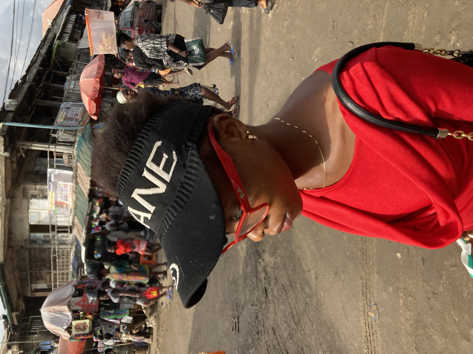 A woman wearing a red top, black sun visor with white lettering, and red eyeglasses stands on a street in Ura Nta Umuarandu, Nigeria. She is accessorized with a gold chain necklace and a black bag strap over her shoulder. A crowded market scene unfolds in the background, with stalls, merchandise, and people moving along the street. Buildings line the street, and an umbrella is visible among the crowd. The street is paved, showing tire marks.