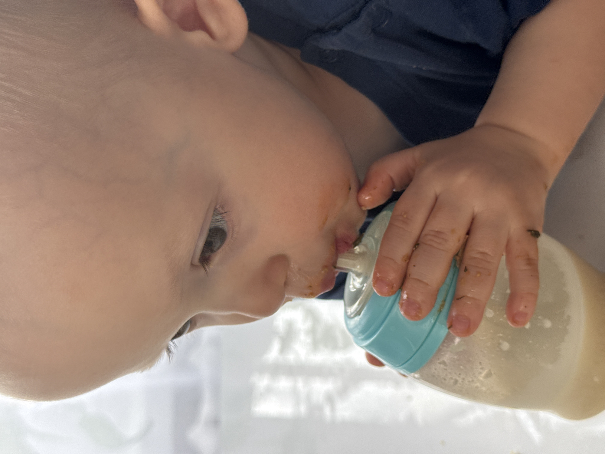 A baby is drinking from a bottle. The baby has fair skin and is wearing a dark blue shirt. The bottle is light blue and clear, filled with a light tan liquid. The baby's hand is holding the bottle. The location and city of Chaikovsky, Russia are not discernible from the image.