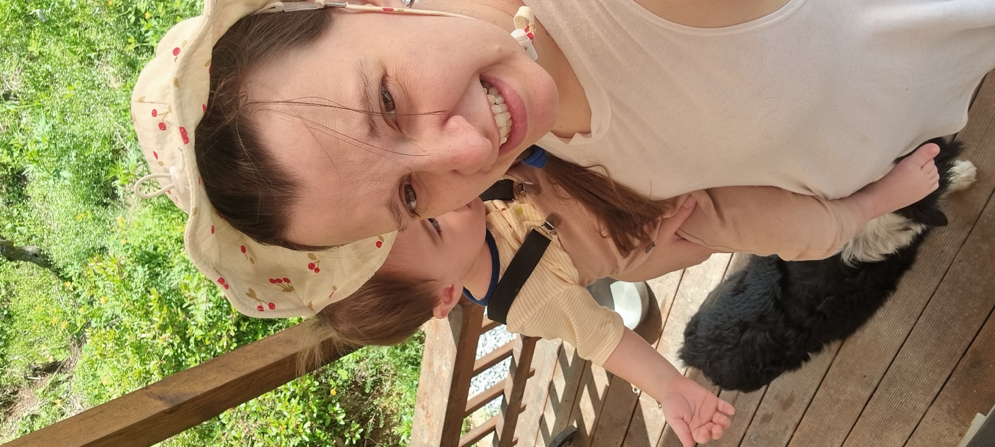 Outside on a wooden deck, a woman smiles while holding a baby. The woman wears a white shirt and a cream-colored sun hat patterned with red cherries. The baby wears a beige outfit with black suspenders and a blue collar. The background shows green foliage. A black and white dog is visible near the woman's hand. The scene is located in Dunnae-myeon, South Korea.