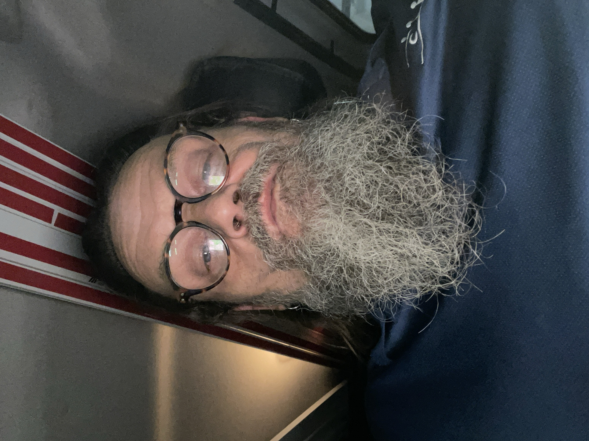 A man with a long, grey beard and tortoiseshell glasses looks directly at the camera. He is wearing a dark blue polo shirt. Behind him, a portion of what appears to be an American flag with red and white stripes is visible. The background is indistinct, but suggests an indoor setting, possibly a vehicle or a room with muted lighting. The image was taken in Raanana, Israel.