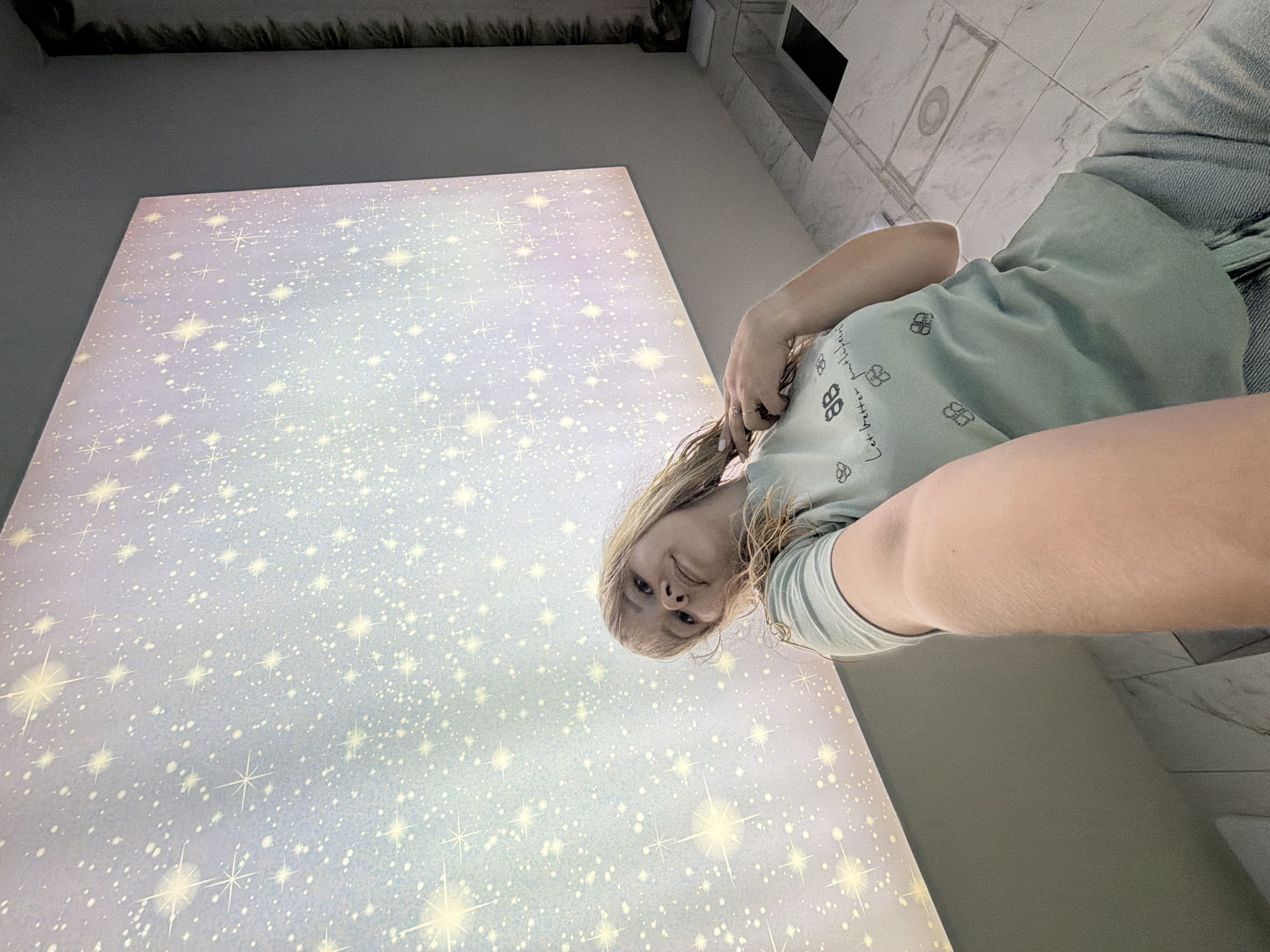 An individual, appearing to be female, is captured in a low-angle self-portrait in Chaikovsky, Russia. She has light-colored, shoulder-length hair and fair skin. Her head is slightly tilted, and she looks directly upward into the camera lens. Her right arm (viewer's left) extends towards the camera, suggesting she is holding the device. Her left hand (viewer's right) is positioned near her neck, with visible rings on her fingers. She wears a light green t-shirt adorned with black graphics, including a stylized "BB" logo, the text "Let better participate," and small clover or butterfly shapes. Light blue pants or jeans are visible from the waist down.
The immediate background is dominated by a large, rectangular, backlit panel mounted on a light gray wall. This panel depicts a simulated starry night sky, featuring numerous small white dots and larger, multi-pointed starbursts, with a subtle light blue to pinkish gradient. The floor is composed of polished white marble-effect tiles, which feature a dark, rectangular floor recess and several tiled grates. To the far right, a dark, textured vertical element, possibly decorative foliage, runs along the wall, above a small recessed shelf containing an unidentified dark object. A light-colored counter-like surface is also visible on the right.