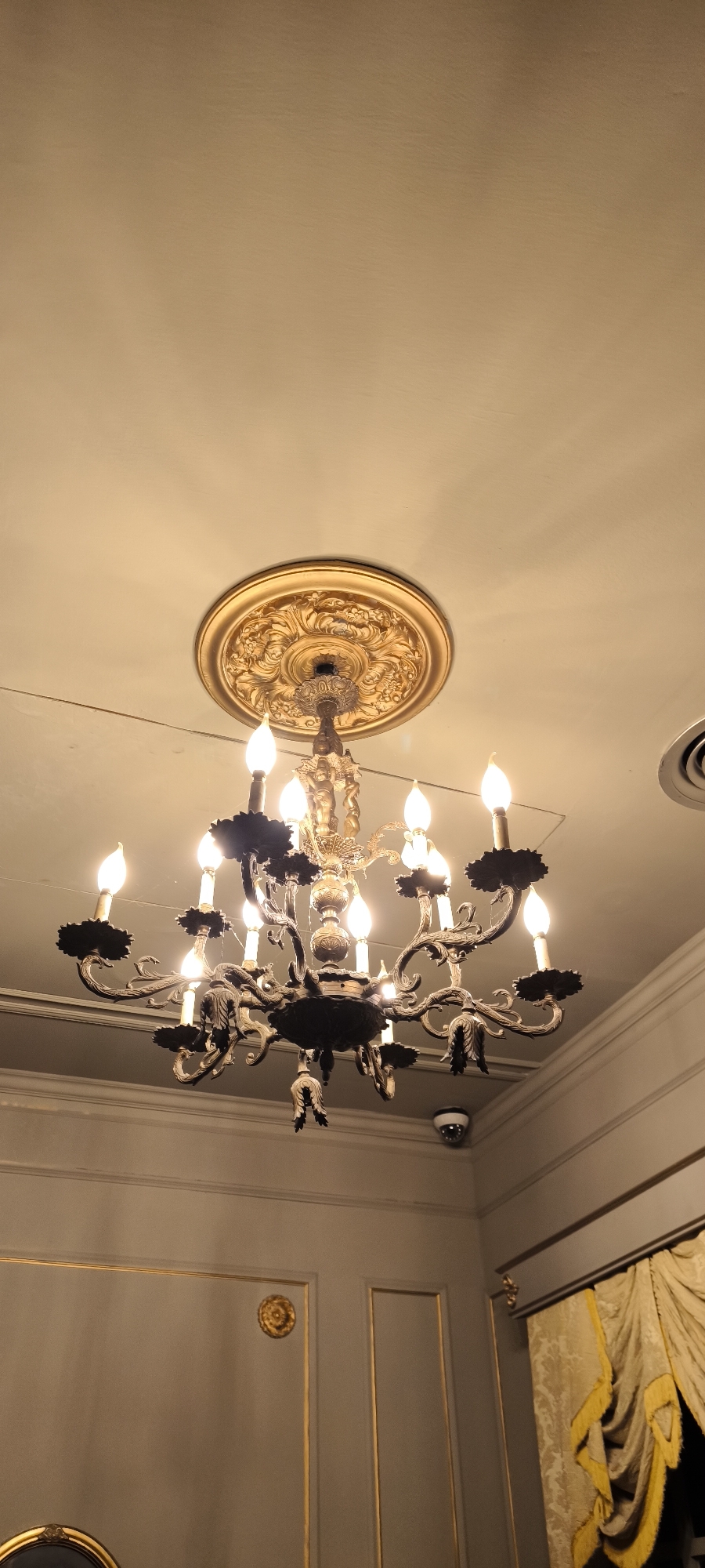 The image depicts an ornate, multi-tiered chandelier hanging from a decorative gold medallion on a white ceiling. The chandelier is silver and black, with multiple faux-candle bulbs emitting a warm light. Below the chandelier, the walls are paneled in gray with gold trim. In the corner of the room, a yellow and gold curtain is visible. A surveillance camera is mounted on the wall, partially obscured by the chandelier. A partial view of a round, dark object is visible in the lower left corner. The location is likely an interior space in Barcelona, Spain, based on the architectural style and decor.