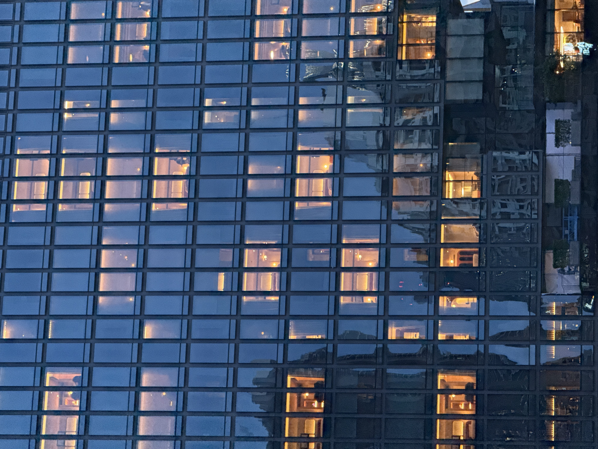 This is a mid-range exterior shot of a modern skyscraper in Bangkok, Thailand. The building is characterized by its grid-like facade of numerous glass windows, most of which are illuminated from within with a warm, orange glow, suggesting occupancy. The time appears to be dusk or early evening, given the deep blue of the sky and the artificial lights within the building.
The windows are arranged in a regular pattern across multiple stories. In some windows, faint silhouettes of people can be discerned, engaged in activities such as sitting at desks or standing. Other windows reveal office furniture, partitions, and lighting fixtures.
Reflections are visible on the glass surfaces, indicating the presence of other buildings or structures in the vicinity, though these are indistinct. The overall impression is one of a bustling urban environment where work continues after dark. The building's architecture is sleek and contemporary, a common style in major metropolitan areas like Bangkok.