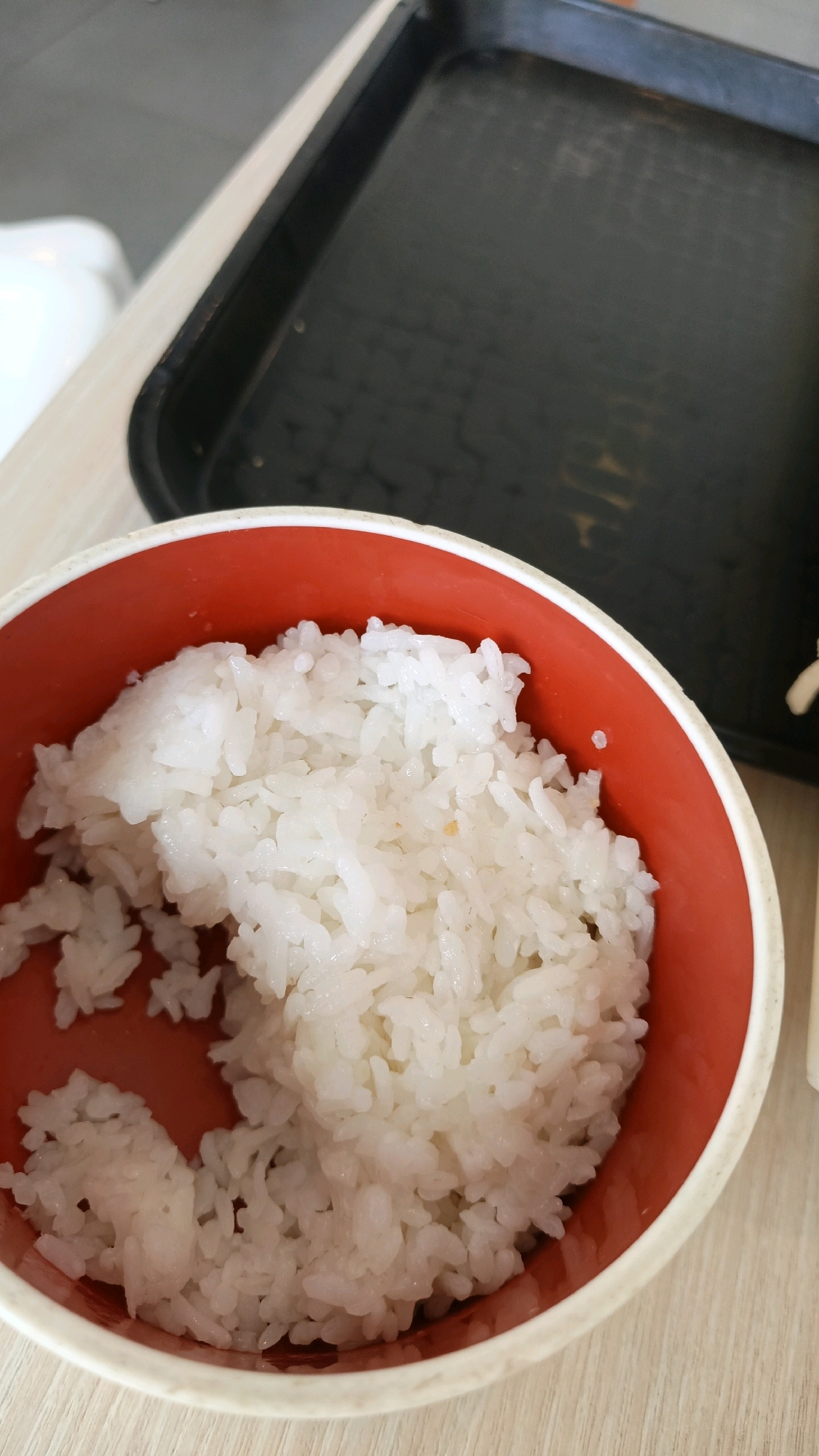A close-up, high-angle shot shows a red and cream-colored bowl filled with white rice, placed on a light-colored wooden surface. A black plastic tray is visible in the background, out of focus. The lighting appears to be from above, casting soft shadows. The setting is likely a restaurant or dining area in Karawang, Indonesia. No people are visible in the image, and no actions or interactions are depicted.