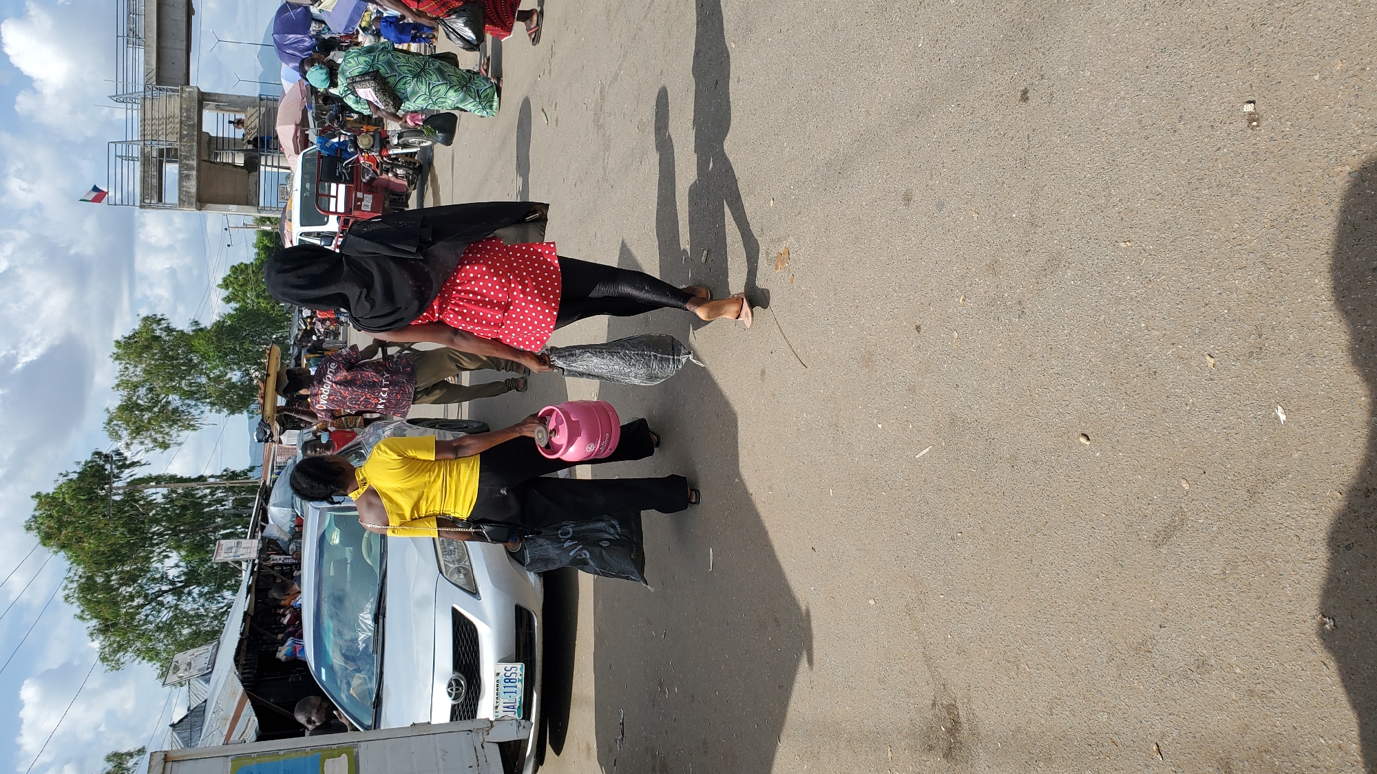 Two women walk away from the camera on a paved road in Jalingo, Nigeria. The woman on the left wears a yellow off-the-shoulder top and black pants, carrying a pink gas cylinder and a black plastic bag. The woman on the right wears a red and white polka dot dress over black leggings, carrying a large black plastic bag. In the background, a silver sedan is parked, and a market with various stalls and people is visible. Several trees are scattered throughout the scene, and the sky is partly cloudy. A pedestrian overpass is located in the upper right corner.