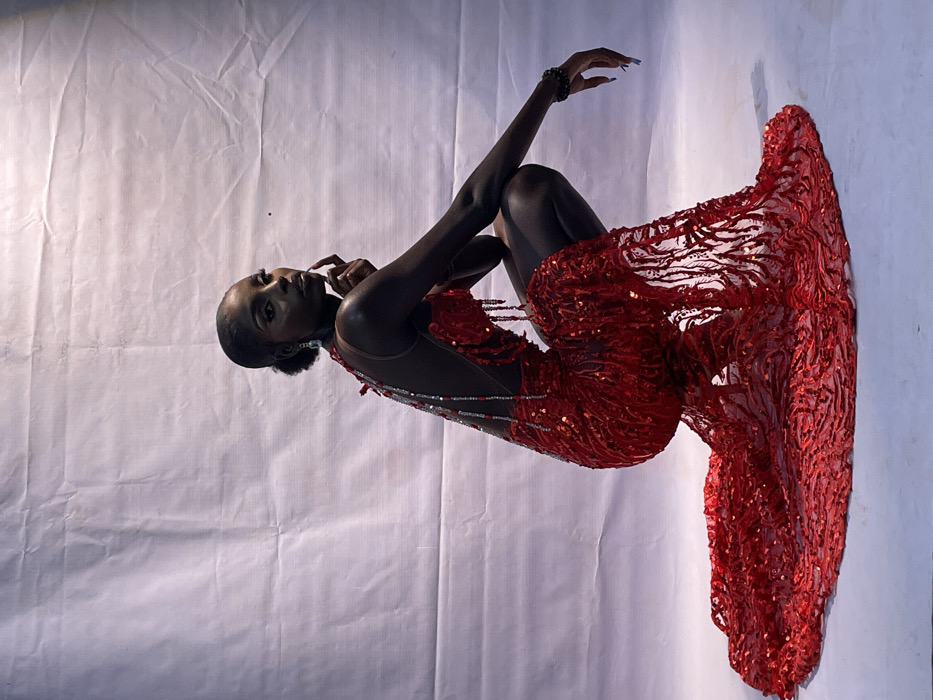 A slender, dark-skinned woman in a striking red gown poses against a white backdrop. She is seated with her knees pulled up and her arms gracefully curved, one hand touching her chin. The dress is intricately embellished with sequins and beads, featuring a flowing, layered skirt. Her hair is styled back, and she wears a beaded bracelet. The image is rotated clockwise with noticeable lighting irregularities across the backdrop.