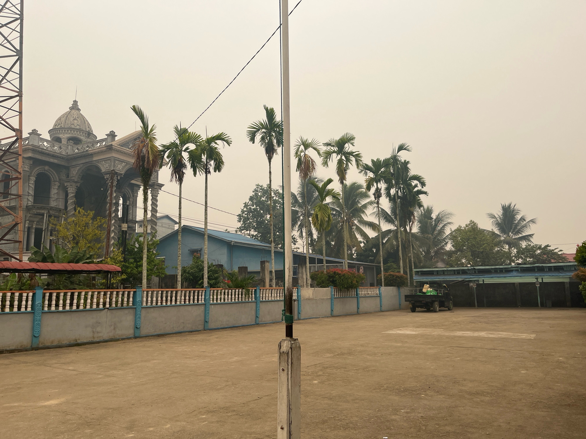 The image depicts an overcast day in Meliau Hulu, Indonesia. In the foreground, a dirt lot is visible with a white pole standing center frame. To the left, a gray fence with blue accents and white balusters runs along the property line. Beyond the fence, a large, ornate, grey building with a domed roof stands prominently, juxtaposed with a simpler, light blue building behind it. Palm trees are scattered throughout the scene, with several lining the fence and taller ones visible behind the buildings. In the mid-ground, a small black truck is parked facing left, carrying what appears to be green and white bags. The background is filled with more palm trees and the hazy sky, suggesting potential smoke or fog.
