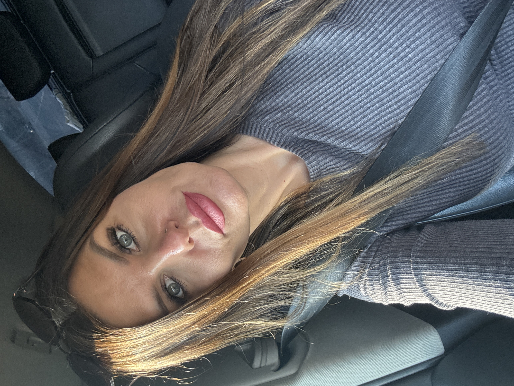 A woman is in the driver's seat of a car, looking into the camera. She has long, brown hair with blonde highlights. Her eyes are green, and she is wearing makeup, including pink lipstick. She is wearing a grey, ribbed sweater and a seatbelt is across her chest. Sunglasses are resting on top of her head. The interior of the car is visible, including black leather seats and a grey dashboard. The background is blurred. The location of this image is not discernible from the visual information provided.