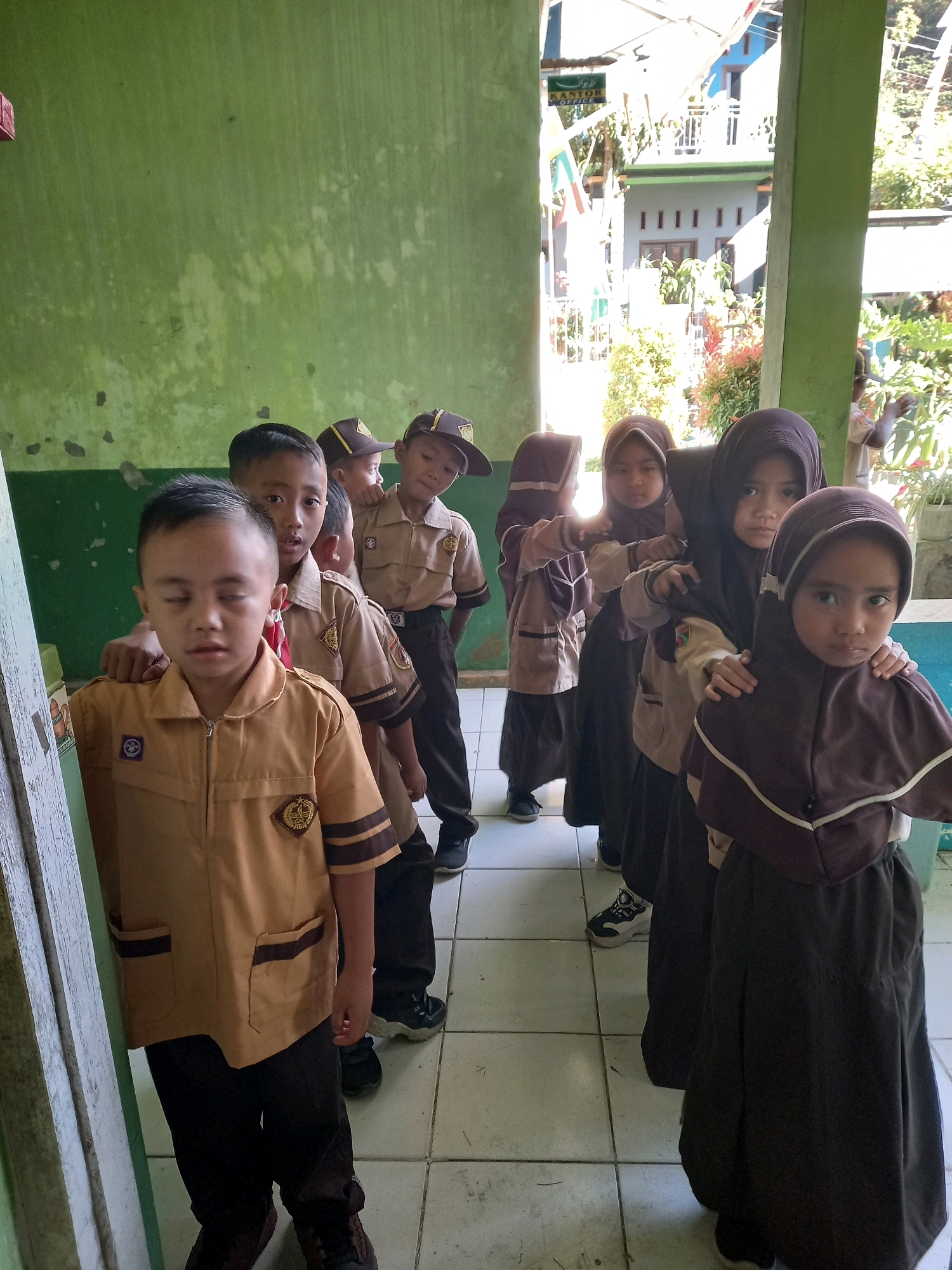 A group of young Indonesian children, likely students, are standing in a line on a tiled floor indoors. The children are wearing school uniforms. The boys in the front wear tan shirts with brown trim and matching hats. The girls behind them wear dark brown hijabs and skirts, with lighter tan tops. The child in the foreground is a boy, facing slightly away from the camera with his eyes closed. The children appear to be waiting or posing for a photograph. The environment appears to be a school hallway or covered walkway, with a green wall on the left and a view of outdoor foliage and buildings through an opening on the right. A sign with Arabic script is visible on the right side. The lighting suggests it is daytime.