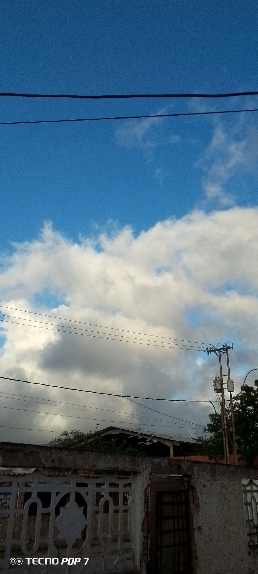 The image depicts a cloudy sky above a residential area. In the foreground, there is a white decorative fence with a metal gate. Behind the fence, a building with a dark roof is visible, partially obscured by trees. Utility poles with electrical wires crisscross the sky. The bottom left corner of the image displays a watermark indicating "TECNO POP 7." The scene appears to be located in Altagracia, Venezuela.