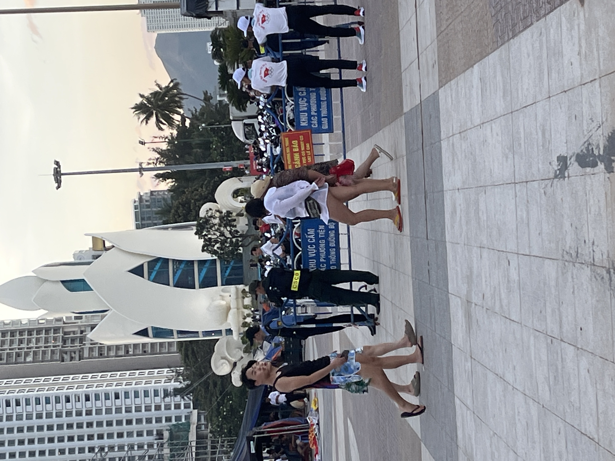 In Nha Trang, Vietnam, at sunset, a crowd gathers in front of a large, white, lotus-shaped structure, possibly a monument or landmark. Two men, one wearing a black tank top and patterned shorts, the other in a similar outfit, walk on the paved plaza. Behind them, two individuals, likely security personnel, stand near a blue barrier with Vietnamese text, one in a dark uniform with "CSCO" visible on their vest, the other in a blue shirt and dark pants. Further back, several people in white shirts and caps are also present, some facing away from the camera. The background features tall buildings, palm trees, and a hazy mountain range, indicative of the coastal city's urban and natural landscape.