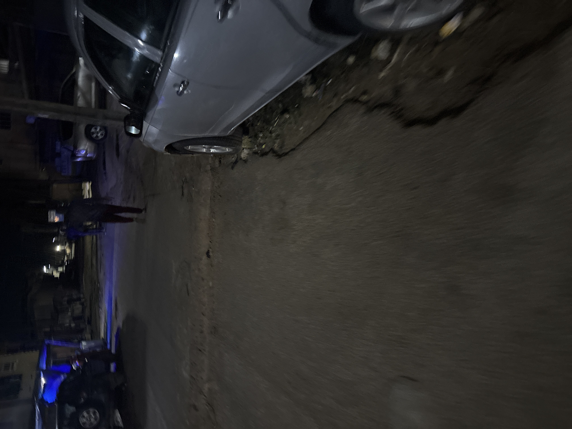 The image depicts a nighttime street scene in Onitsha, Nigeria. A silver sedan is parked on the right side of the frame, its side and wheel visible. In the background, a dark SUV with a blue light is parked in front of a building. A person wearing dark clothing and red pants is walking away from the camera towards the left of the frame. The street appears to be unpaved or in disrepair, with visible potholes and debris. Streetlights are present in the background, casting a dim illumination on the scene. The overall impression is one of a moderately lit urban environment at night.