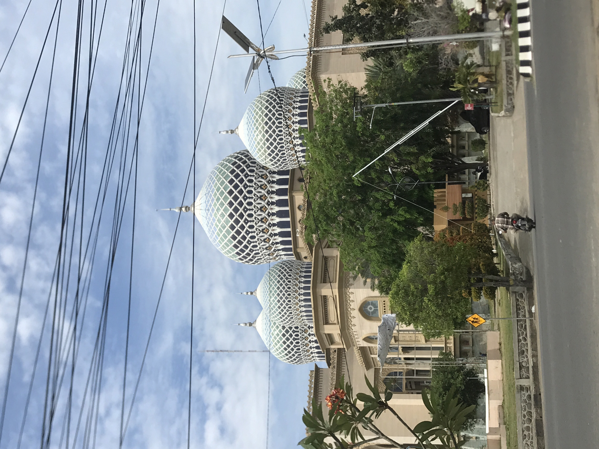 The image shows the Baiturrahman Mosque in Lhokseumawe, Indonesia, viewed from street level. The mosque features three prominent domes with a geometric lattice pattern. Power lines are visible in the foreground against a cloudy blue sky. A street runs along the right side of the image, with a pedestrian crossing sign and a person on a motorbike. Vegetation, including trees and flowering plants, is present in the midground. The architectural details of the mosque's facade are visible, including windows and decorative elements.