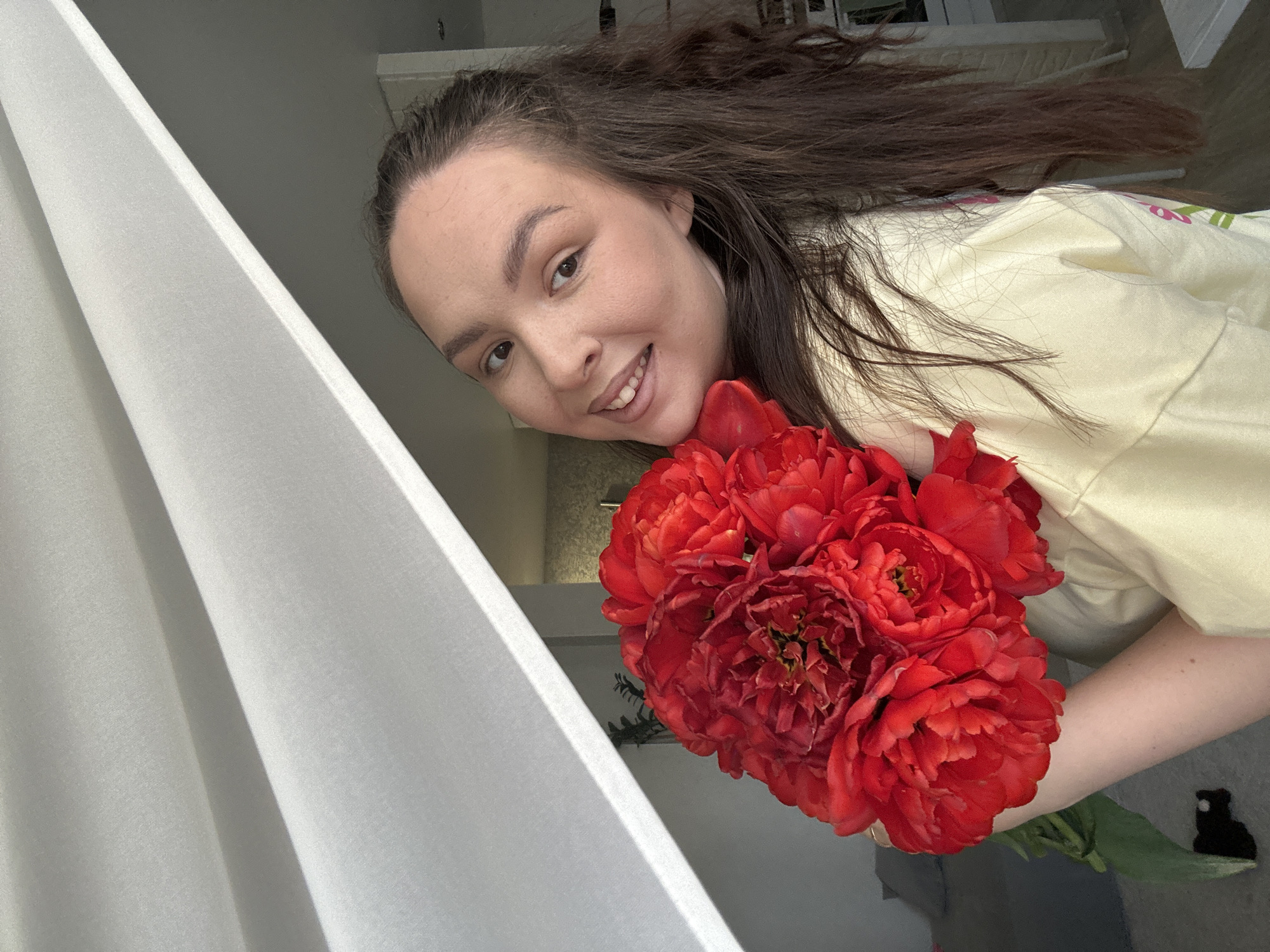 A woman is holding a bouquet of red flowers. She is wearing a light yellow shirt and has long brown hair. A white curtain is to the left of the image. The location is Chaikovsky, Russia.