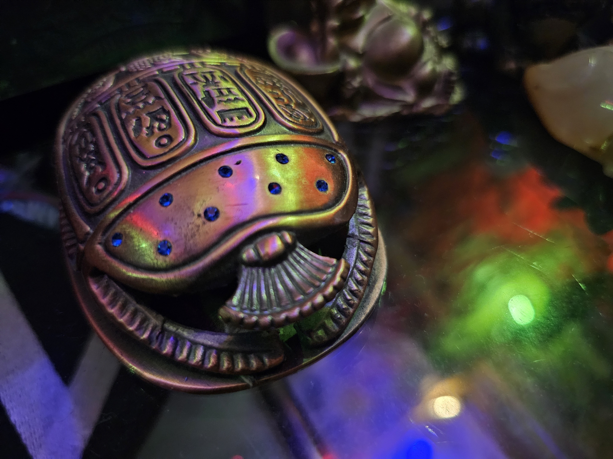 The image features a close-up of an ornate, metallic scarab beetle figurine. The figurine is a copper or bronze color and is decorated with what appear to be hieroglyphic symbols etched into its carapace. Several small, dark blue, jewel-like accents are embedded in the front of the scarab. The figurine is placed on a reflective surface, possibly glass, which displays a colorful and blurry background of lights in shades of purple, green, red, and blue. The lighting suggests a dark environment, possibly indoors with artificial lighting. The focus is sharp on the scarab, with the background lights creating a bokeh effect. There are no people visible. The overall mood is somewhat mysterious due to the lighting and the ancient Egyptian motif of the scarab. No specific time of day, weather, or location cues are evident, beyond the implied indoor setting. The visible text consists of stylized symbols on the scarab's shell, resembling Egyptian hieroglyphs, but they are not clearly legible.