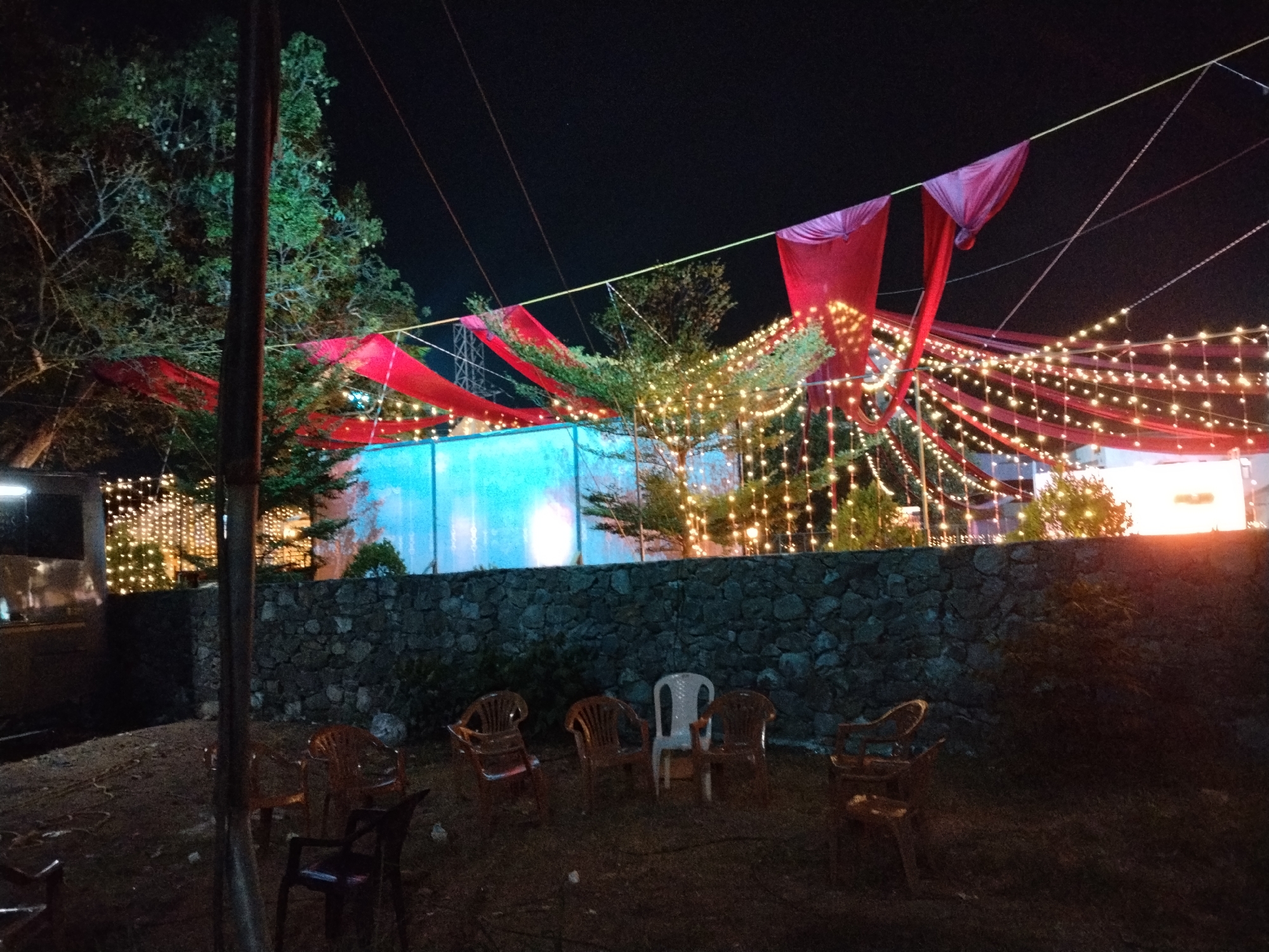This is an outdoor night scene depicting a festive or event setup in Kalamassery, India. The setting is illuminated by numerous warm-toned string lights (fairy lights) draped across trees and overhead, creating a vibrant and celebratory atmosphere against the dark night sky.
Overhead, red fabric drapes are strung between points, contributing to the decorative theme. Large trees with dense foliage are visible, particularly on the left and in the background, with many branches adorned with the glowing lights. In the mid-ground, a rough-hewn stone wall provides a boundary, behind which a translucent light-blue structure or screen is brightly lit, adding a cool contrast to the warm lights.
In the foreground, several plastic chairs in various shades of brown, white, and black are scattered on what appears to be a dirt or grassy ground, suggesting a casual seating area for guests. A dark, thick vertical pole or tree trunk stands prominently on the left side of the frame. The overall impression is one of a lively evening gathering or event, characterized by its enchanting lighting and simple yet effective decorations. No people are visible, and there is no discernible text in the image.