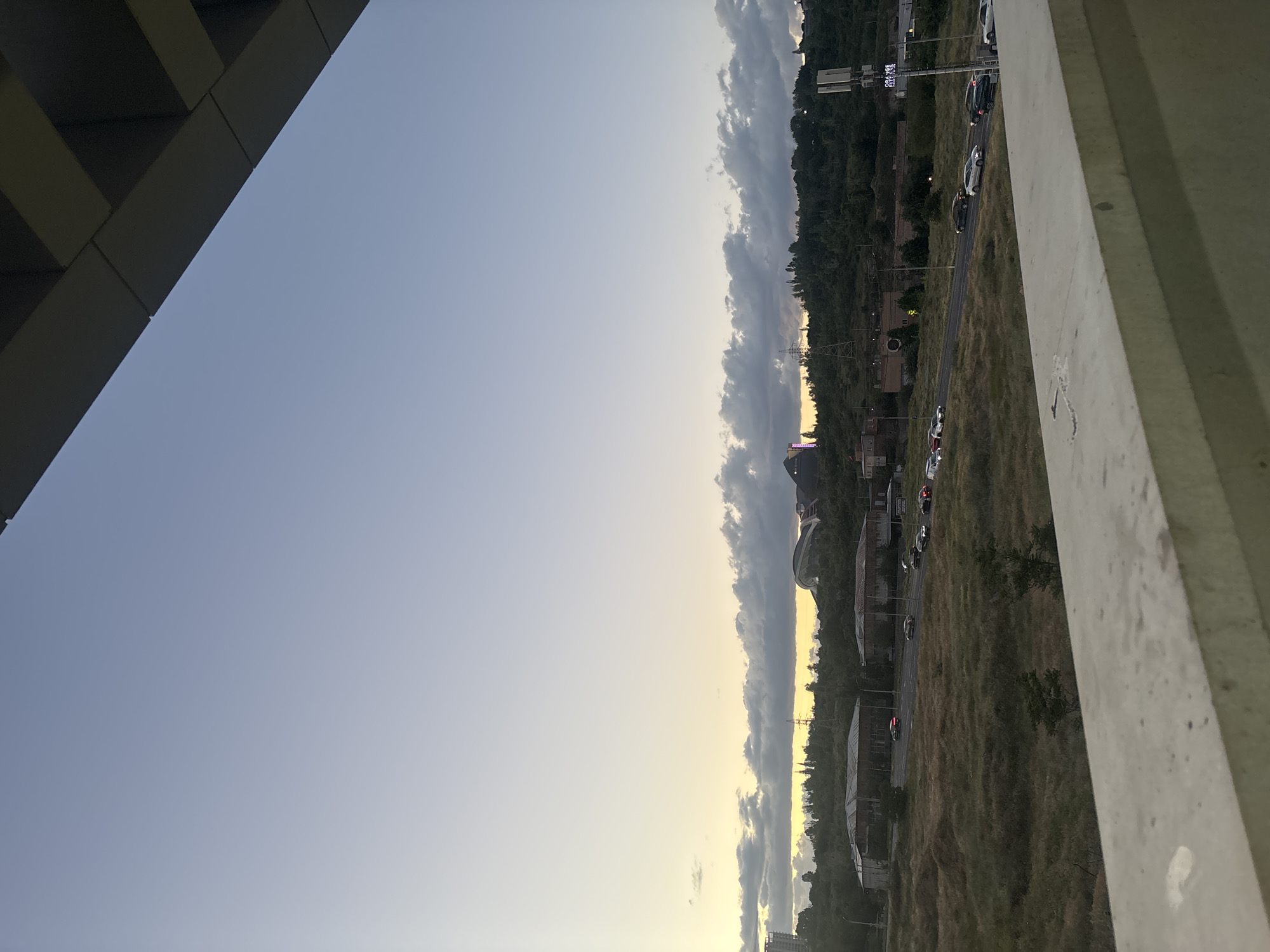 This image, captured from an elevated perspective, displays an urban or semi-urban landscape under the warm glow of either sunrise or sunset. The scene is dominated by a vast sky that transitions from a pale yellow-orange at the horizon to a clear light blue higher up. A prominent band of dark, elongated clouds stretches horizontally across the mid-sky, silhouetted against the brighter light from the sun.
In the foreground, an architectural element, likely a part of a building or balcony, cuts diagonally across the upper left corner, providing the vantage point for the shot. Below the horizon, a road snakes through the landscape, populated with cars that show visible headlights and taillights, suggesting ongoing traffic. The terrain surrounding the road is characterized by dry, grassy fields.
Further into the midground and background, the landscape includes scattered buildings, utility poles with power lines, and distant hills or mountains densely covered with trees. On the right side, large modern buildings with distinctive curved roofs are visible, potentially part of a stadium or a large complex. One of the hint crops reveals a digital display in the distance, possibly showing numbers like "17:35". Another faint, vertically oriented sign with unreadable text is also present near the road. The overall lighting and the active car lights strongly suggest the time of day is dusk or early evening. No specific people are identifiable, but the scene is active with vehicles. The mood is tranquil, capturing the transition of light at day's end or start.