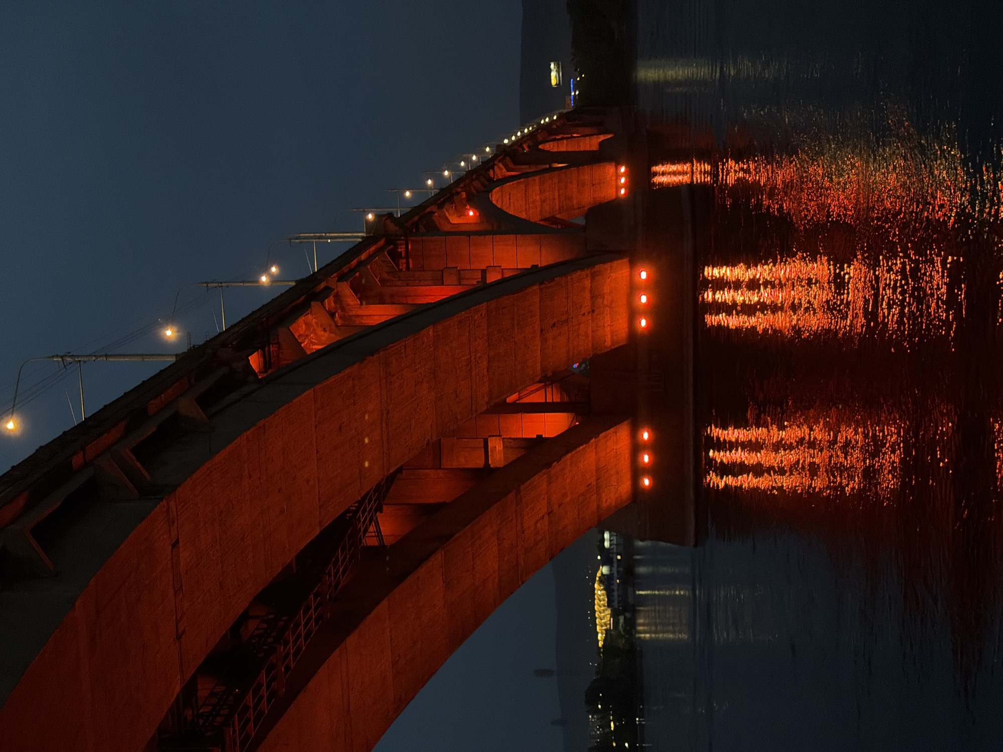 The image shows the Krasnoyarsk Railway Bridge at night, illuminated with red lights. The bridge spans a body of water, likely the Yenisei River, and its red arches are reflected in the dark water. Streetlights are visible along the top of the bridge, and in the distance, faint lights of the city of Krasnoyarsk can be seen. The sky is a deep blue, indicating nighttime. There are no people or animals visible in the image. The scene is industrial and architectural, focusing on the bridge's structure and lighting.
