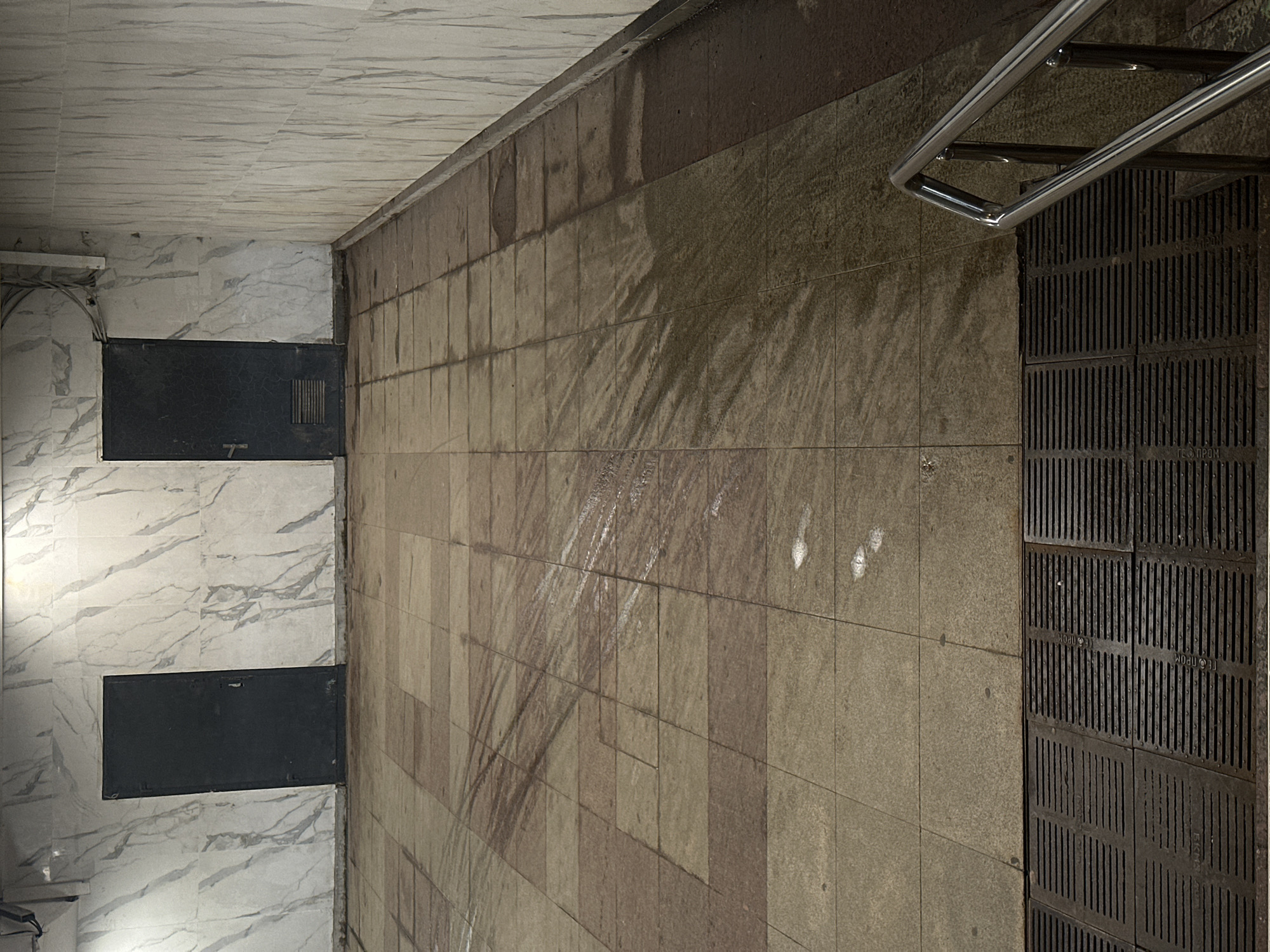The image displays an interior view of what appears to be a public passage, possibly an underpass or a section of a metro station. The scene is artificially lit and devoid of people.
On the left, a wall and the ceiling are covered in white tiles with a grey marble pattern. On this left wall, two rectangular, dark-colored panels are visible, possibly access doors or utility covers. The lower panel has a handle mechanism.
The right wall is constructed from light brown or beige square tiles. This wall shows significant wear and tear, with numerous dark, streaky stains running vertically and diagonally, suggesting water damage or heavy grime accumulation. Several distinct white marks are also scattered across this tiled surface.
The floor is partially covered by these same light brown tiles on the left side, but transitions to a large area of dark metal grates on the right, adjacent to the right wall. These grates feature vertical slats and appear robust, likely for drainage or covering an open area. A shiny, silver-colored metal handrail extends downwards from the upper right portion of the image, positioned above these grates, indicating a nearby staircase or ramp.
Visible text on several of the dark metal grates includes the Cyrillic word "ГЕОПРОМ" (GEOPROM) and the acronym "ЕКНЗ". The overall impression is one of functional, utilitarian public infrastructure, showing signs of use and environmental exposure, consistent with an urban setting like Moscow, Russia.