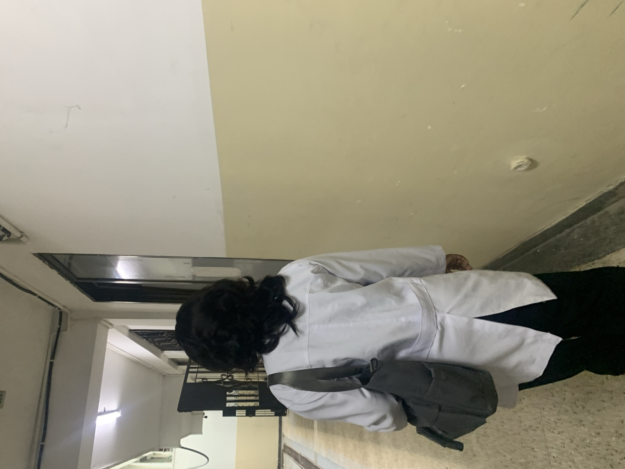 A person is seen from behind, walking down an indoor hallway or corridor. They are wearing a white lab coat over dark clothing and have dark, curly hair. A dark grey shoulder bag is carried across their body.
The setting features two-toned walls, with a white upper section and a light yellow or beige lower section. The floor is light-colored, likely concrete or tile. On the left side, a dark banister or railing suggests a staircase or an upper-level area. Overhead, there are dark, industrial-style ceiling elements, possibly a grate or ventilation system, and a bright fluorescent light fixture is visible further down the hallway.
The person appears to be moving away from the camera. The internal lighting conditions suggest it could be daytime, augmented by artificial light sources. The attire and environment imply a location such as a hospital, clinic, or university building. There is no visible text in the image.