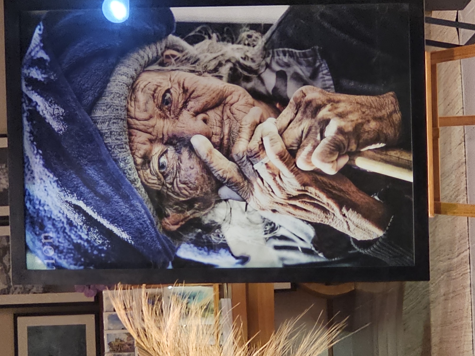 The image is a close-up photograph displayed on a television screen. The photograph features an elderly person with heavily wrinkled skin and gray hair, appearing to be wearing a head covering or shawl, with their hands positioned near their face. The person's face is the main focal point, with the texture of the skin and the details of the hands highlighted. The television screen is framed and positioned above a surface where dried, wheat-like stalks are arranged. A framed picture is visible beneath the television. The environment suggests an indoor setting, possibly a home or gallery. There is no explicit information or visual clues in the photograph to pinpoint the location to Denpasar, Indonesia, or any specific location.