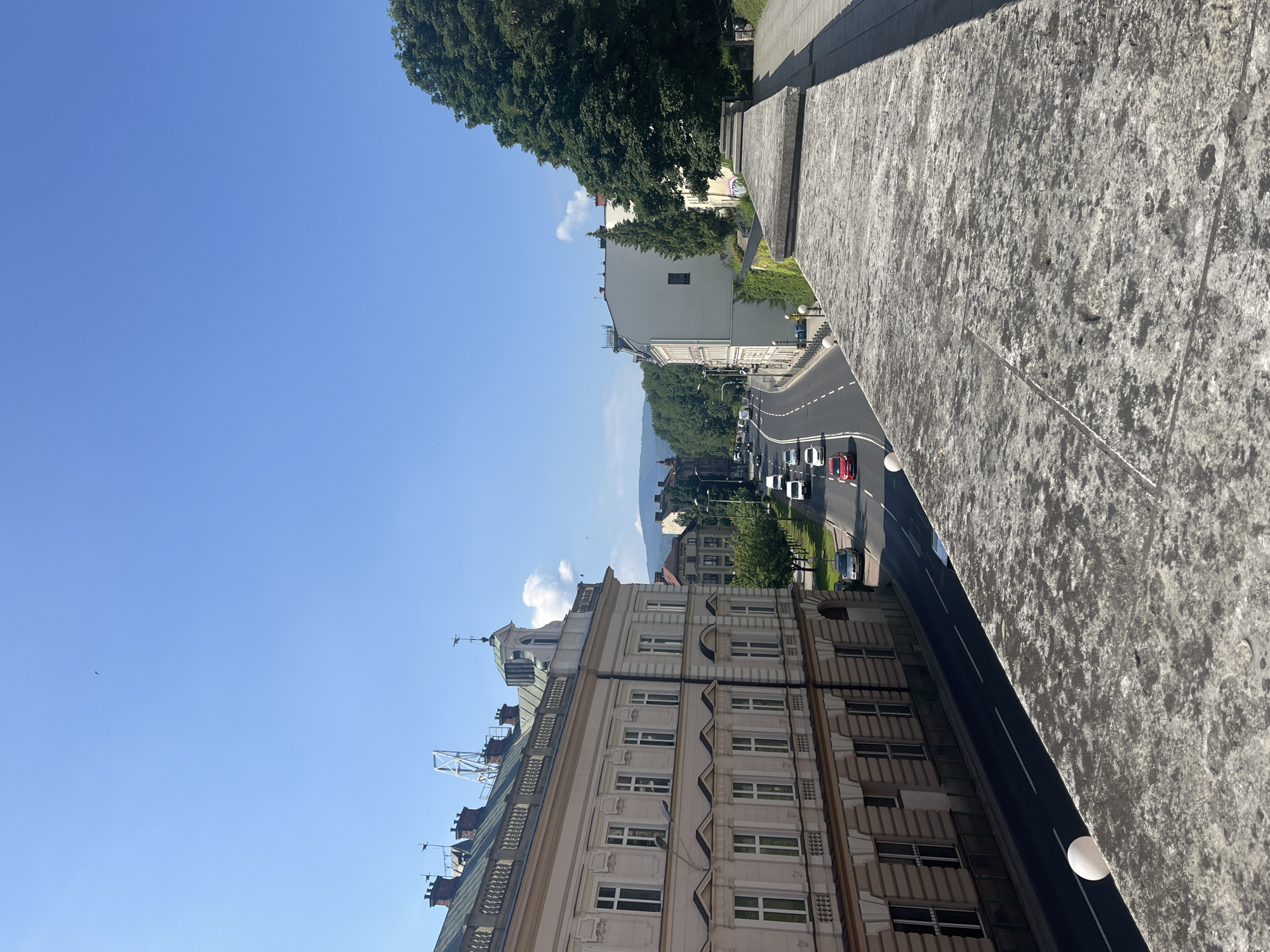 The image shows a street scene in a city with buildings on either side. In the foreground, there is a stone wall with a textured surface. Beyond the wall, a multi-lane road runs through the city, with several cars driving on it. There are trees and other buildings lining the road. The sky is clear and blue, with a few faint clouds visible. The architecture suggests a European city, possibly with a historical center. The overall impression is one of a sunny day in an urban environment.