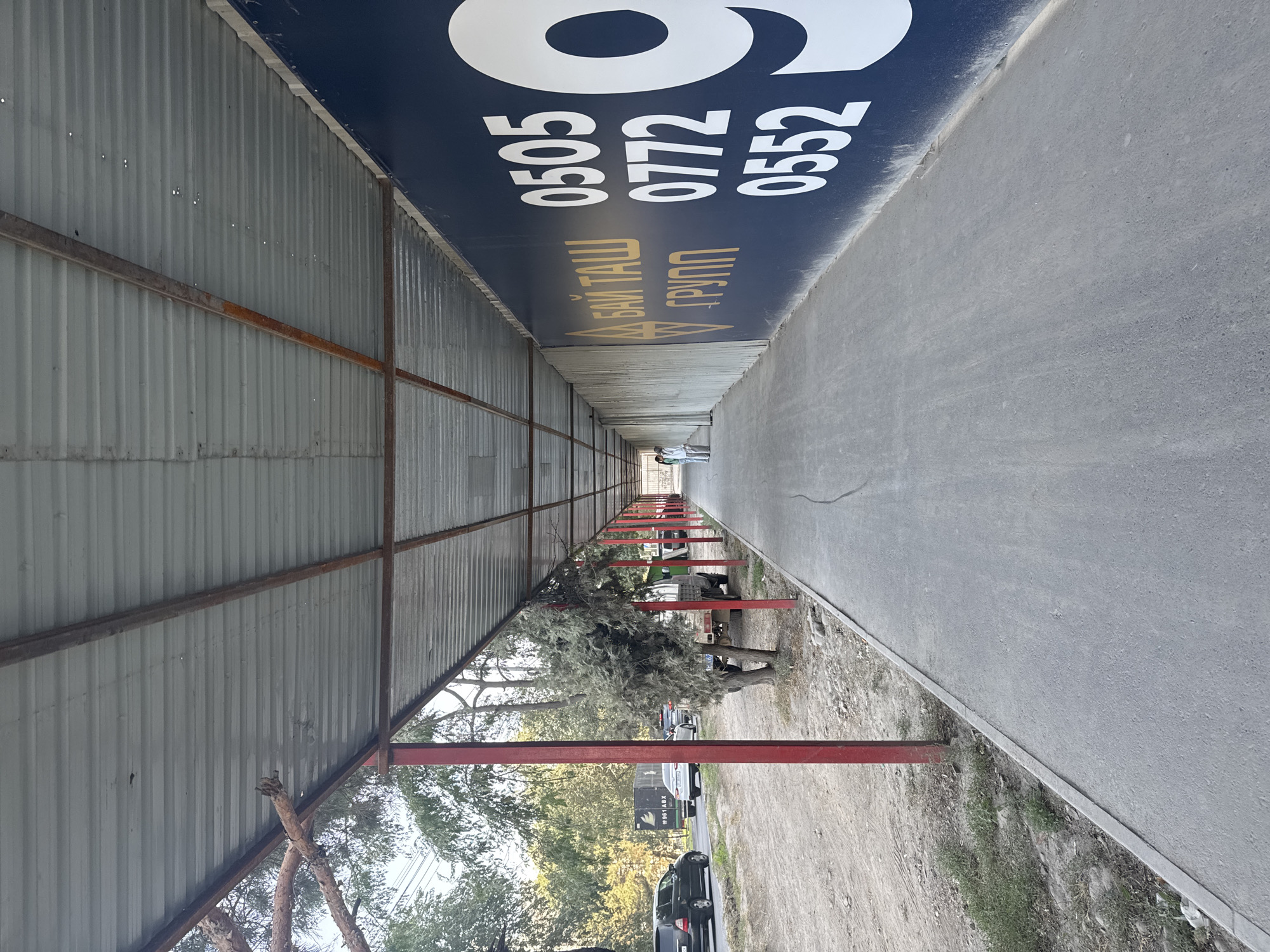 This image depicts a covered walkway or tunnel situated in Bishkek City, Kyrgyzstan. The walkway is constructed with a corrugated metal roof supported by a metal frame, with red metal poles lining the left side. A dark blue banner with gold lettering and white numbers is prominently displayed on the right side of the walkway. The banner features the words "БАЙ ТАШ ГРУПП" in gold and the numbers "0505", "0772", and "0552" in white, along with a large white numeral "9". Two individuals are visible in the distance at the far end of the walkway, appearing to be walking away from the camera. To the left of the walkway, a road with several cars is visible, indicating an urban setting. Trees and foliage line the roadside. The ground within the walkway is paved with concrete, showing some wear and tear. The overall scene suggests a commercial or construction area, possibly an entrance or passage to a development.