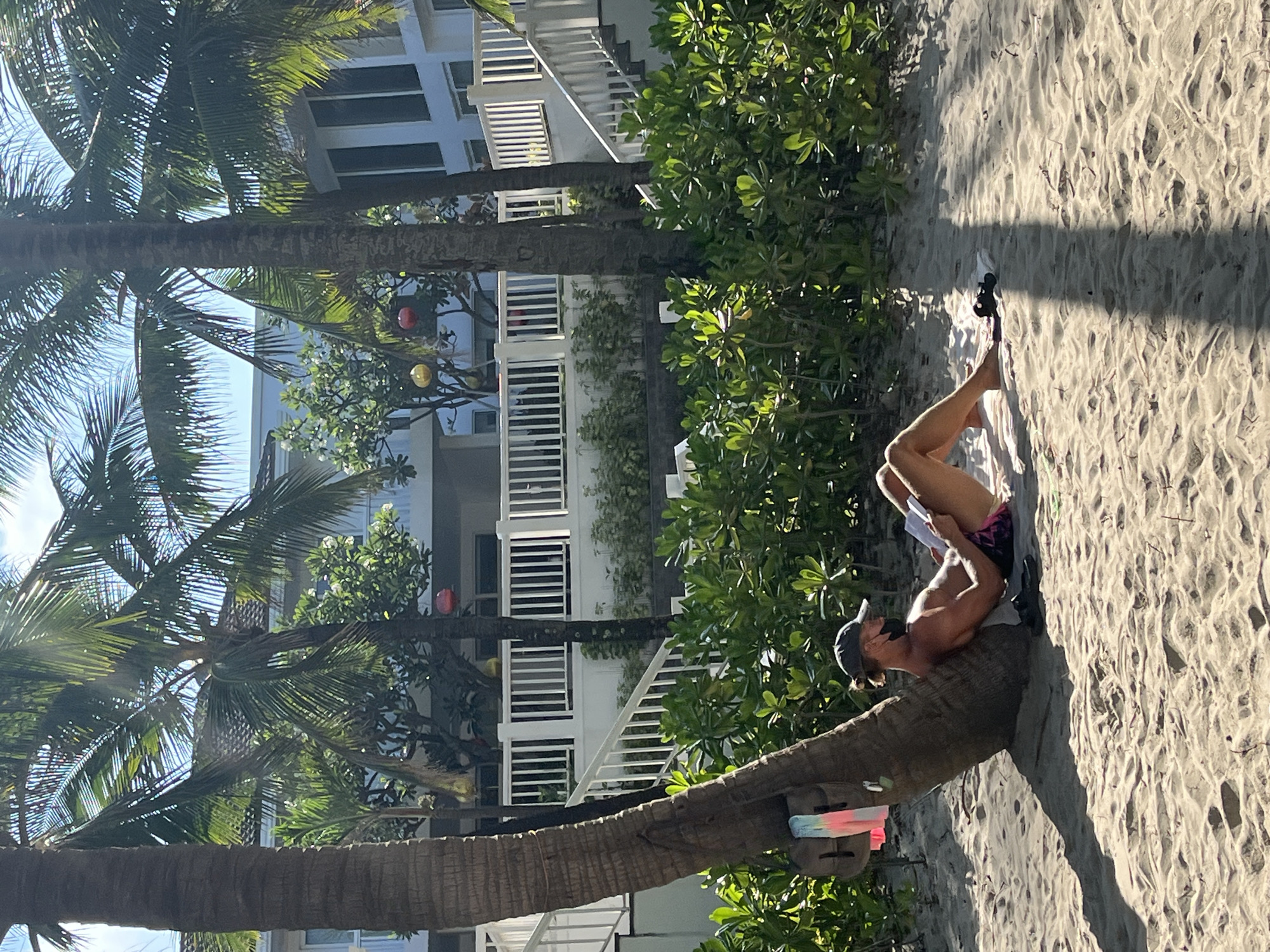 A sunny, outdoor, eye-level shot presents a man reclining on a palm tree trunk at a beach in Đà Nẵng, Vietnam. The man, wearing shorts, a cap, and dark shoes, is reading a document. He is positioned lengthwise along the trunk, on the sandy beach. Palm trees are visible on the left, and a white building with railings can be seen in the background.