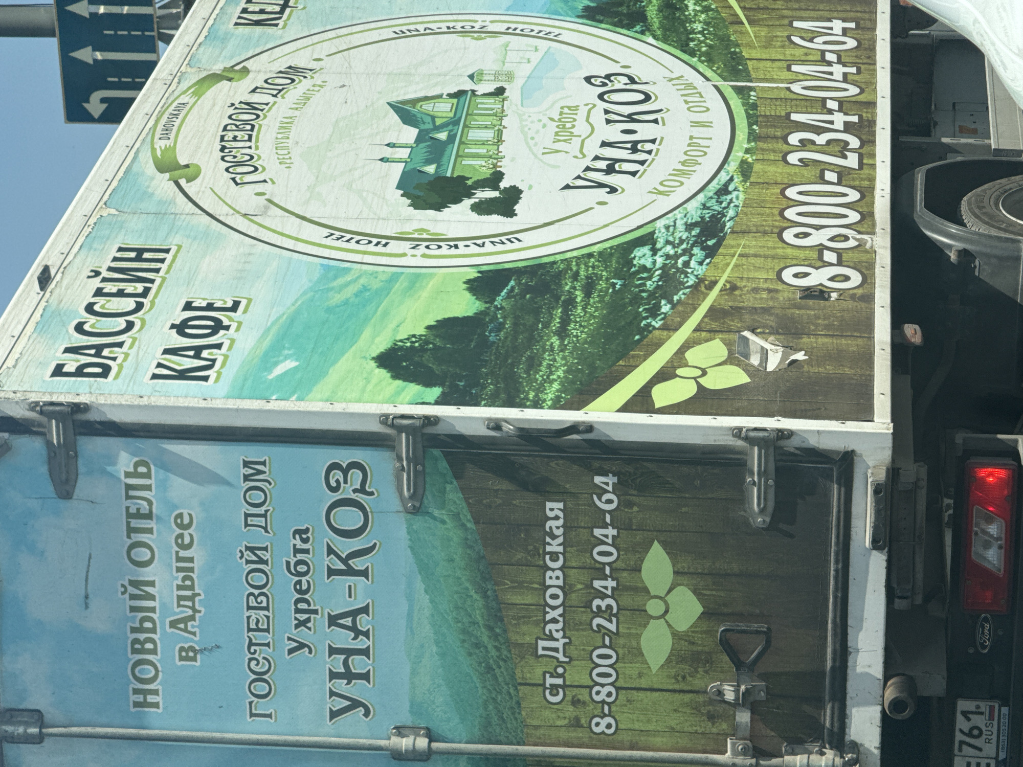The image shows the side of a white truck with a blue and green graphic advertisement. The advertisement features text in Russian. On the left side, it reads "НОВЫЙ ОТЕЛЬ В АДЫГЕЕ" (NEW HOTEL IN ADYGEYA) and "ГОСТЕВОЙ ДОМ У ХРЕБТА УНА-КОЗ" (GUEST HOUSE AT THE RIDGE UNA-KOZ). Below this, it states "ст. Даховская" (settlement Dakhovskaya) and a phone number "8-800-234-04-64".
The right side of the advertisement features a circular logo with text that includes "ГОСТЕВОЙ ДОМ" (GUEST HOUSE) and "УНА-КОЗ" (UNA-KOZ). The logo depicts a green house with a pool and surrounding trees. It also includes the phrase "КОМФОРТ И ОТДЫХ" (COMFORT AND REST).
The background of the truck's advertisement shows a mountainous landscape with green trees and fields. The truck itself is parked on a road, with a partial view of another vehicle to the right and a traffic sign with arrows pointing left and up in the background. The lower portion of the truck shows a license plate that appears to be from Russia. The graphic is partially worn in some areas.
The image is a photograph of the side of a truck. The lighting suggests it is daytime. There are no people visible in the image. The location of Aksay, Russia is not explicitly visible in the image, but the license plate suggests it is in Russia.