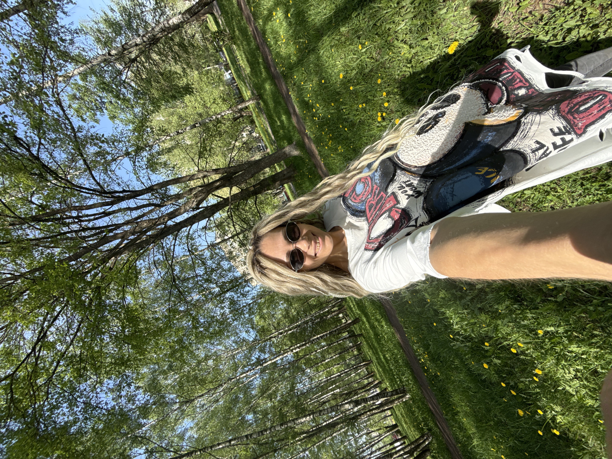 The image depicts a woman in sunglasses lying on a grassy field dotted with yellow flowers. She is wearing a white t-shirt with a graphic print and has long blonde hair. The background shows a line of trees with green leaves, partially obscuring a blue sky. The location is identified as Rusanovo, Russia.