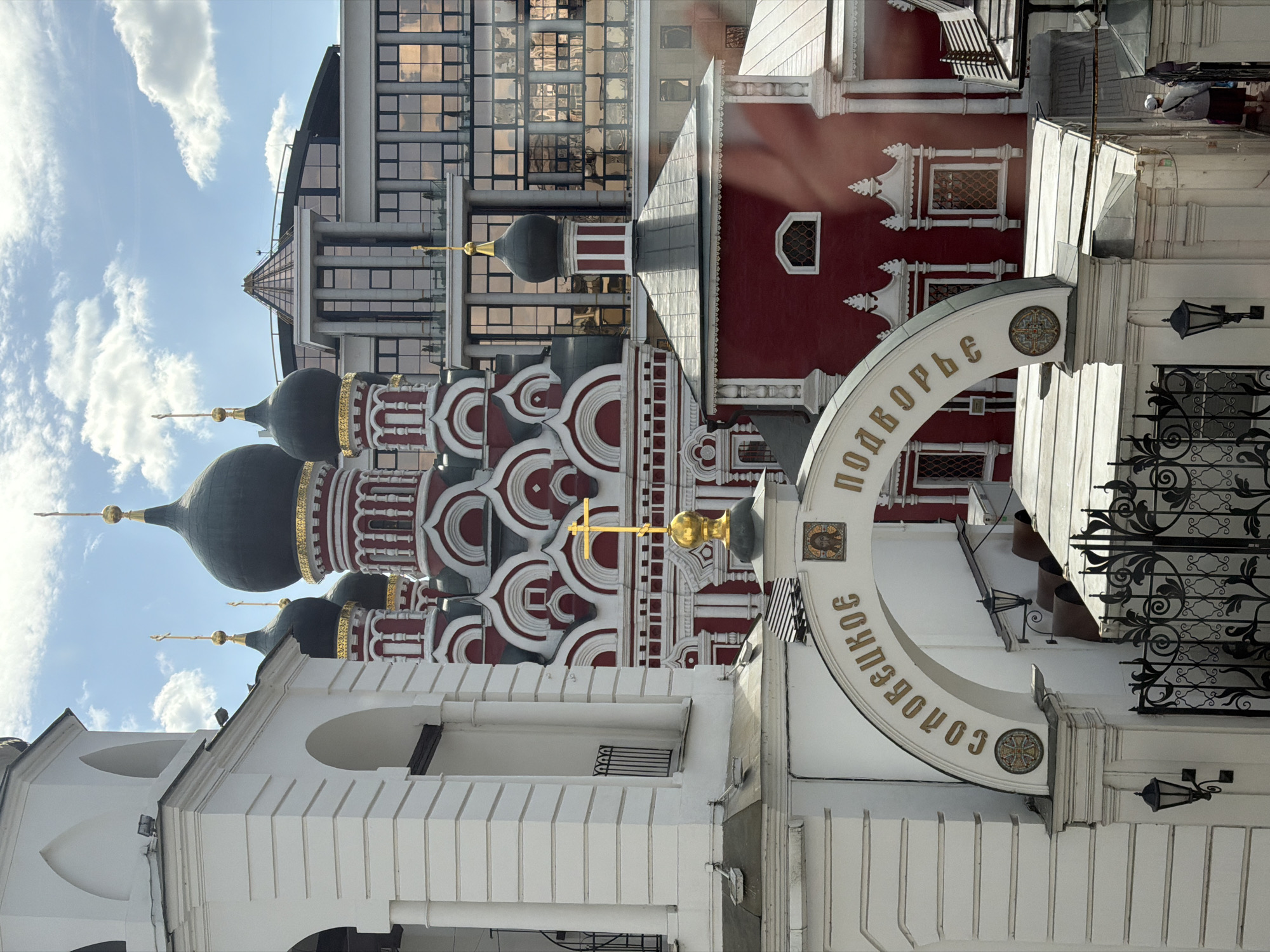 The image shows the Podvor'e of the Trinity-St. Sergius Lavra in Moscow, Russia. The architecture includes the distinctive onion domes and red and white patterned facades of traditional Russian Orthodox churches. A white archway with Cyrillic lettering, marking the entrance to the Podvor'e, is visible. In the background, a modern building with mirrored windows contrasts with the historic architecture of the church. The sky is blue with some clouds. The image is taken from a slightly low angle.