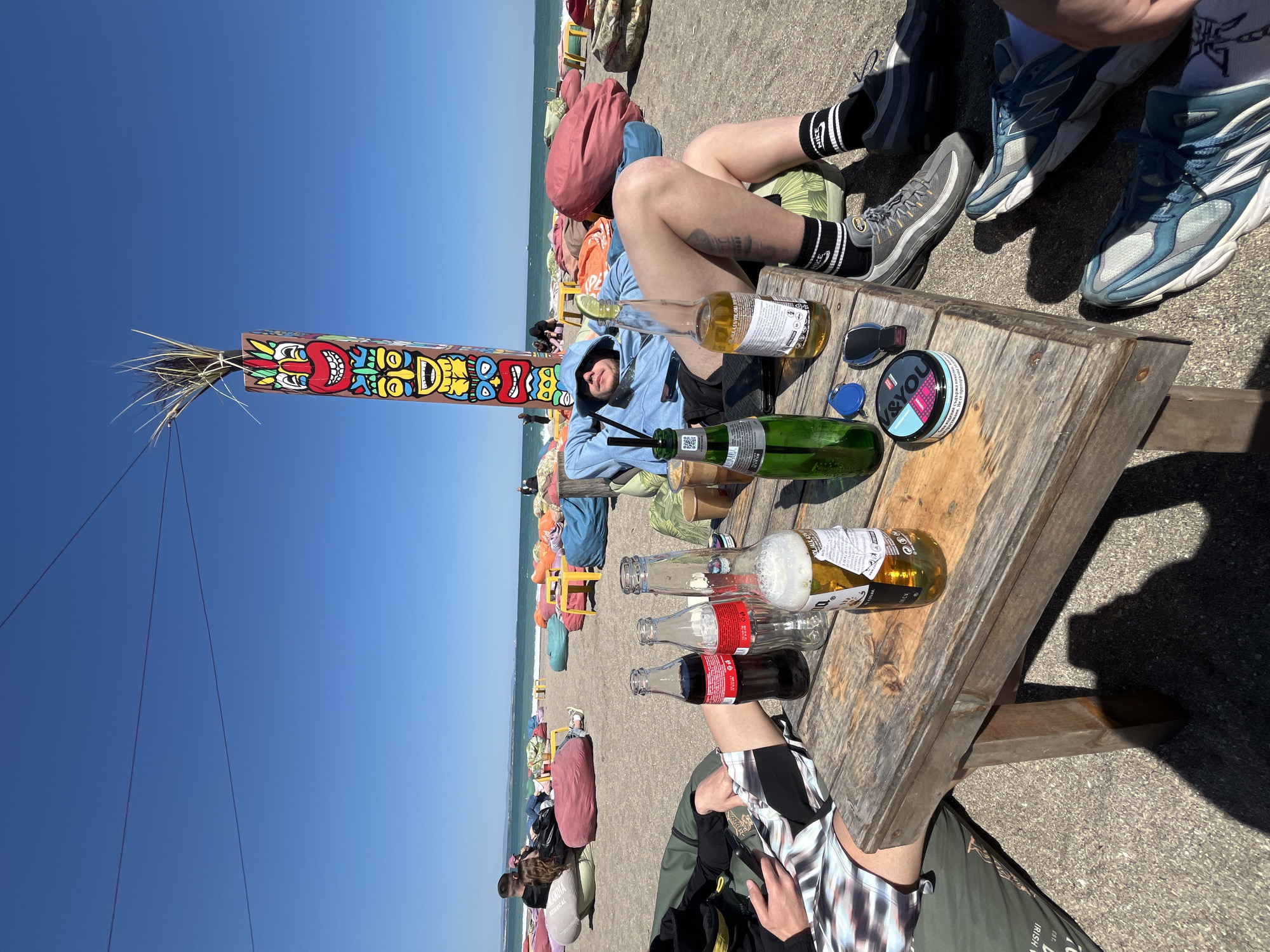 The image depicts a beach scene in Burgas, Bulgaria, under a clear blue sky. In the foreground, a weathered wooden table holds several empty bottles of Coca-Cola and beer, a green glass bottle with straws, a tin of snus, and a pair of sunglasses. Two individuals are positioned around the table. One person wears a blue hoodie and sits amongst colorful beanbag chairs on the sand, while another is partially visible wearing a black and white shirt. To the left of the frame stands a tall, decorated totem pole. In the background, the Black Sea is visible, along with more beachgoers relaxing on beanbag chairs.