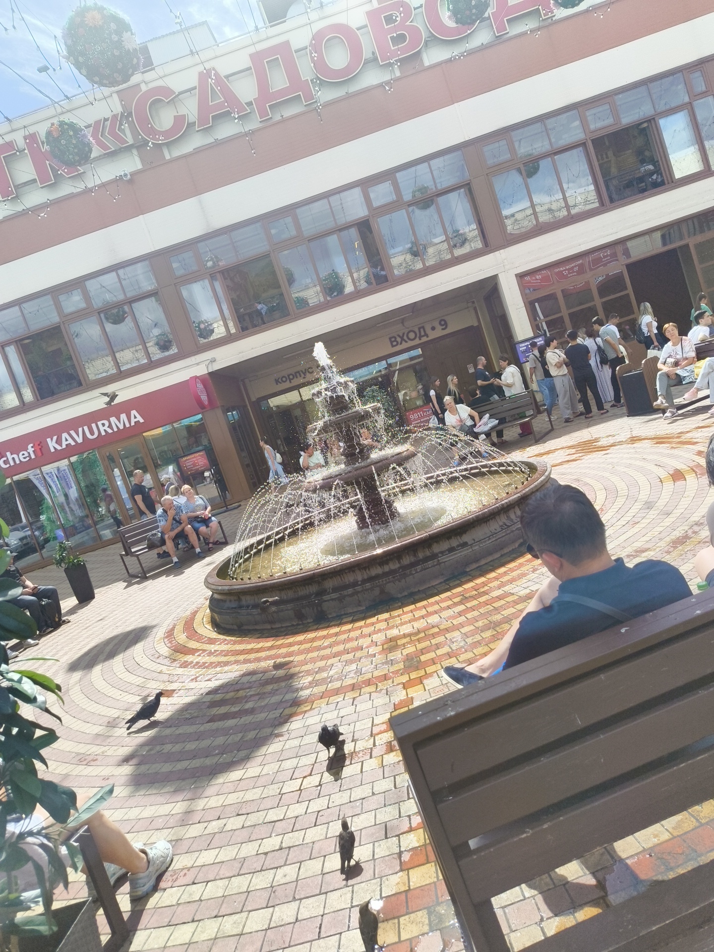The image depicts a public outdoor space with a multi-tiered fountain in the foreground. A man is seated on a wooden bench, facing away from the camera, with his back to the fountain. The background features a building with large windows and signage that reads "САДОВОДС". The building appears to be a shopping center or commercial establishment. Several people are visible in front of the building and near the fountain, some of whom are sitting on benches. Pigeons are present on the tiled ground. The environment suggests a sunny day in a city, likely a pedestrian area associated with a shopping district or a park. The signage is in Russian, indicating a location within Russia. The overall scene is one of a bustling public area on a clear day.