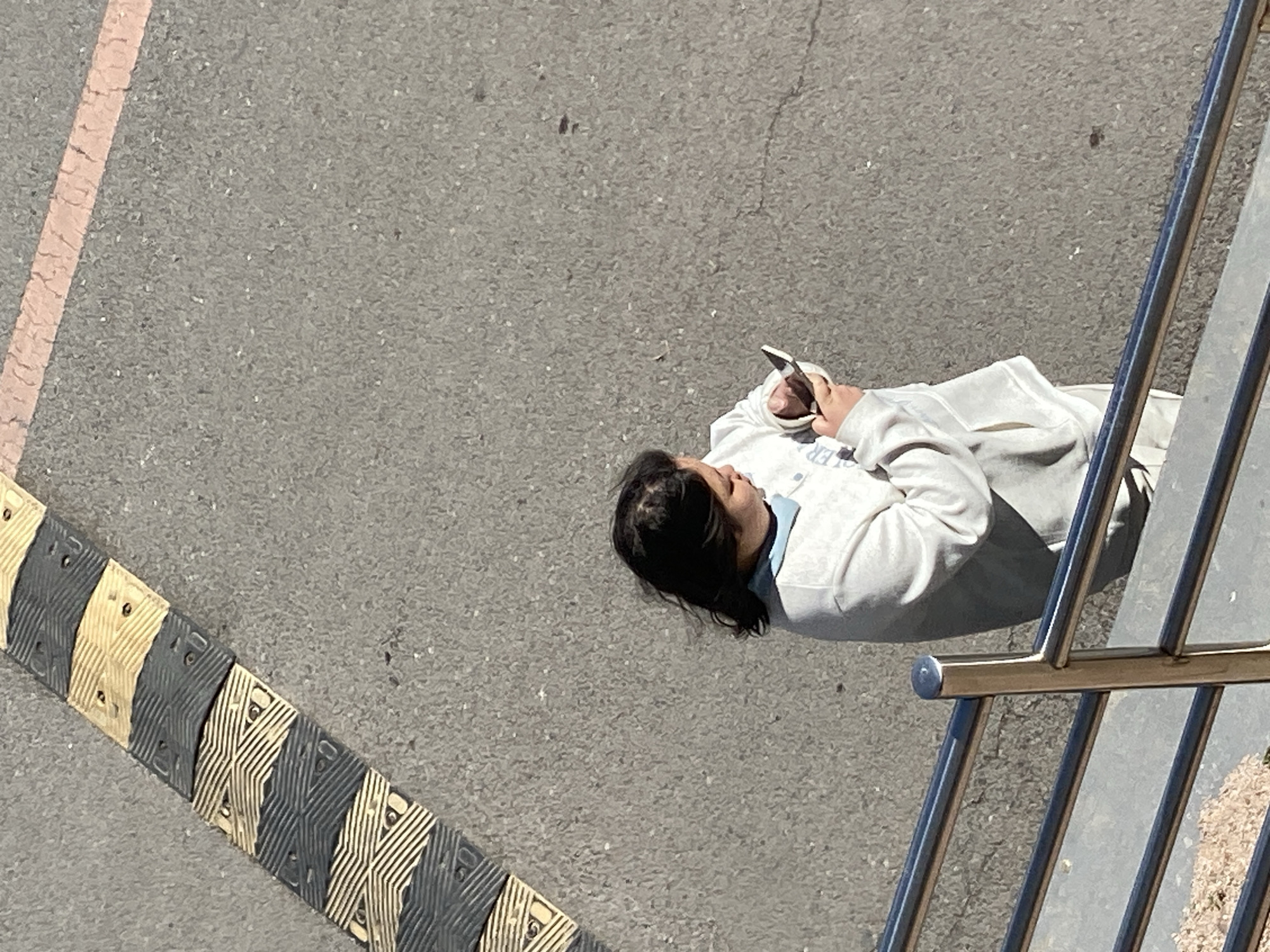 A top-down view shows a person standing on a gray asphalt surface, holding a mobile phone. The individual has dark hair and is wearing a light-colored sweatshirt or jacket. A speed bump with alternating black and light tan stripes is visible on the left side of the frame. A metal railing with vertical bars occupies the right side of the image. The asphalt surface is marked with a pink line on the upper left. The location is consistent with an urban environment in Zhongtun, China, based on the presence of street furniture and asphalt surfaces. The person appears to be using their phone while standing near the railing and speed bump.