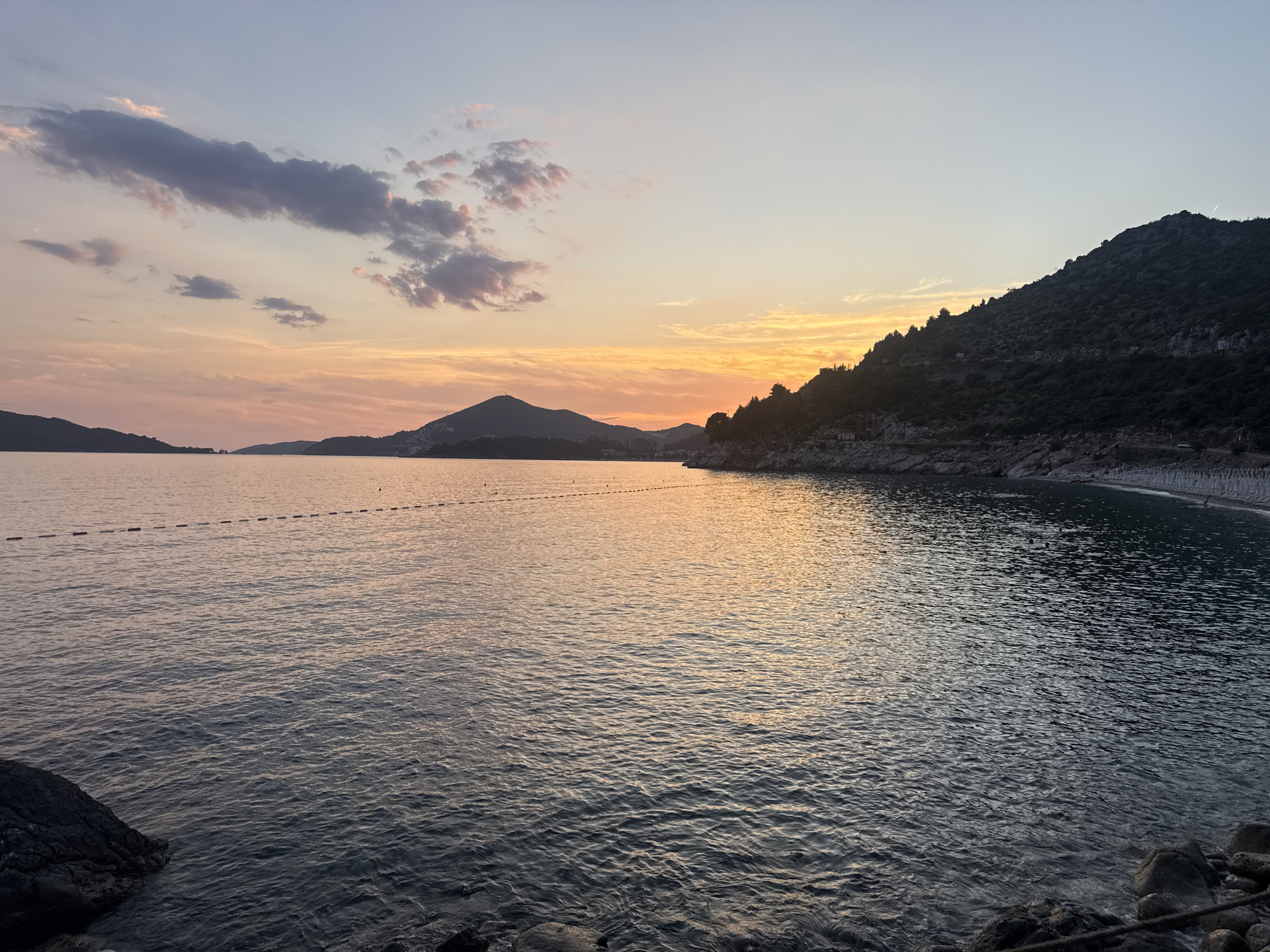 The image depicts a serene sunset scene at Kamenovo beach in Montenegro. The sky is a canvas of soft pastel colors, with hues of orange, pink, and light blue blending seamlessly. Wispy clouds are scattered across the sky, some tinged with the warm glow of the setting sun. In the distance, the silhouettes of mountains rise against the horizon, their dark forms contrasting with the vibrant sky.
The calm sea stretches out before the viewer, its surface gently rippled by a light breeze. The water reflects the colors of the sky, creating a shimmering, iridescent effect. A line of buoys, marked by small, reddish-orange floats, stretches across the water, indicating a designated swimming area or boundary.
The foreground of the image is occupied by a rocky shoreline, with dark, rough-hewn stones scattered along the edge of the water. These rocks provide a grounded, natural element to the scene. The beach itself is visible in the background on the right, appearing as a gently curving stretch of sand or pebbles nestled against the base of a dark, tree-covered hill. The hill slopes down towards the water, its dense foliage creating a rich, dark silhouette.
The overall atmosphere of the image is one of tranquility and natural beauty. The fading light of the sunset casts a peaceful glow over the landscape, inviting a sense of calm reflection.