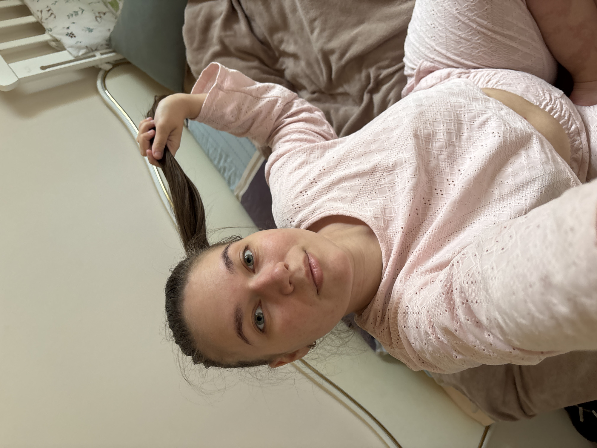 A woman in a pink patterned pajama set is lying on a bed. She has brown hair pulled back in a ponytail and is holding the ponytail up with her hand. The bed has a brown blanket, a blue and gray striped section, and a white railing with a green pillow on top. The background is a plain white wall. The location is Chaikovsky, Russia.