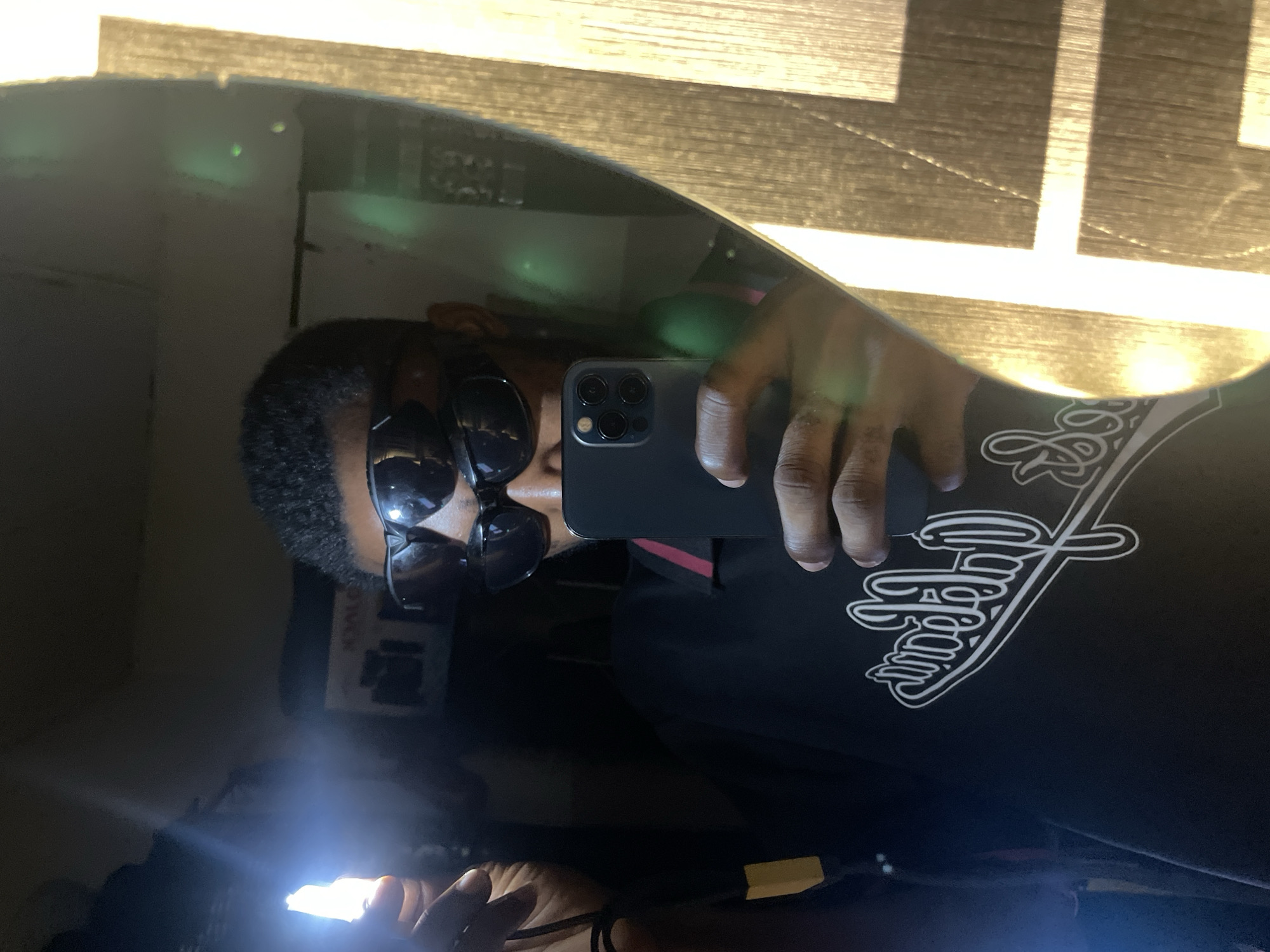 This image captures a mirror selfie taken by an individual in Lokoja, Nigeria. The reflection shows a person, likely male, with dark skin and short, dark hair. The individual is wearing two pairs of dark sunglasses; one pair is positioned over the eyes, and a second pair is resting on the forehead. The person is dressed in a dark polo shirt featuring a red stripe on the collar and a white, stylized script logo on the chest. In their right hand, the person holds a dark smartphone, used to capture the image. The left hand holds a bright, glowing white light source, which emits significant glare and has a visible yellow strap or cord attached. Behind the reflection, a dark interior space is visible, including a partially discernible white box with text and an image of a camera-like device. The right side of the mirror reflects a bright, textured surface, possibly wood, illuminated by a strong light source, creating distinct patterns. Green lens flares are present in the upper right portion of the reflection.