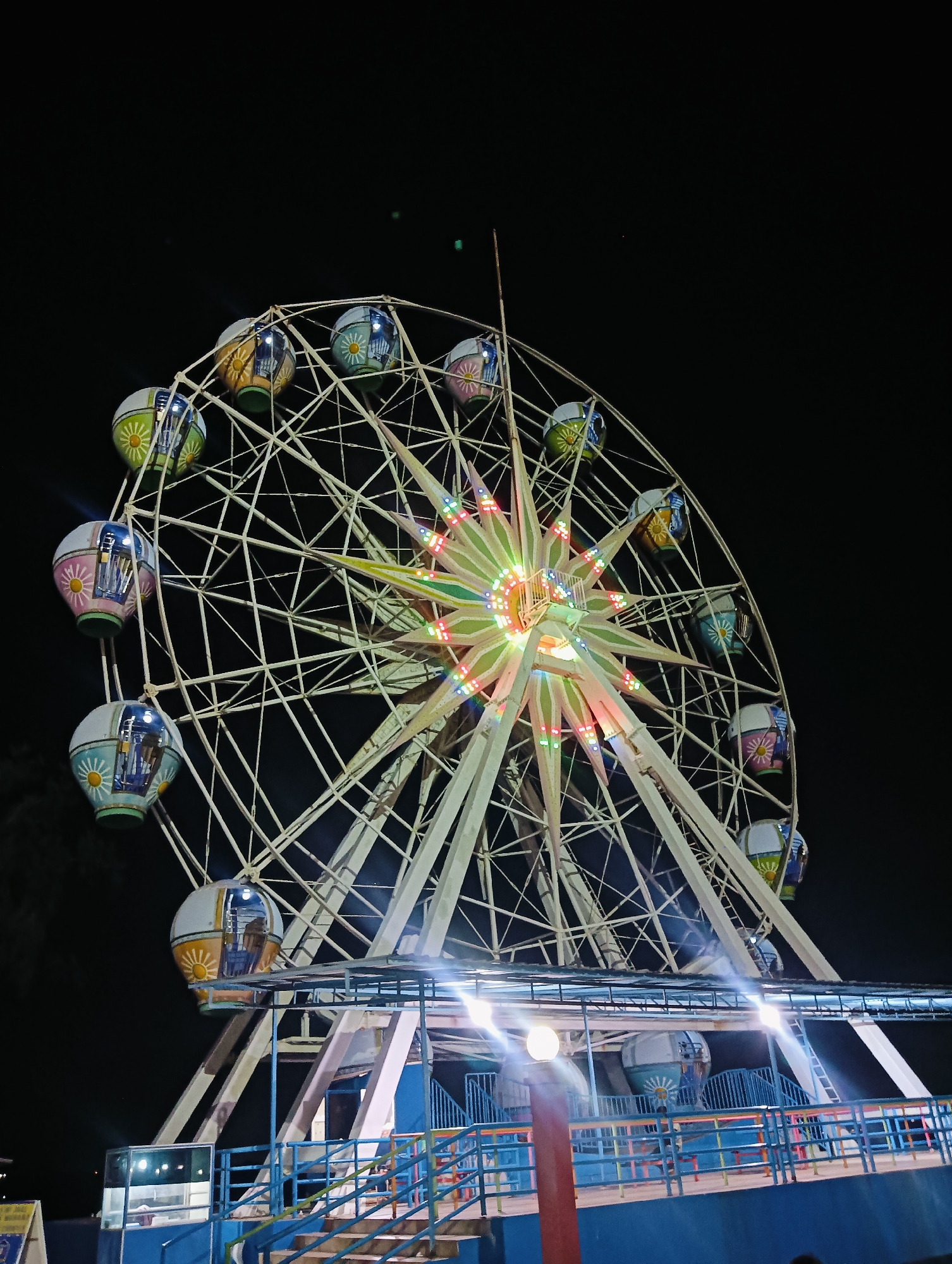 A Ferris wheel stands illuminated against a dark night sky in Batam, Indonesia. The wheel, a light cream color, features a star-shaped center brightly lit with multi-colored bulbs. Its spokes are also illuminated. Dangling from the wheel are numerous gondolas, each painted with a white base and decorated with colorful designs, including floral patterns and sun motifs. The gondolas appear to be occupied by people. At the base of the Ferris wheel is a blue structure with blue railings and stairs, also lit by overhead lights. The overall scene suggests a night-time amusement park or fairground.