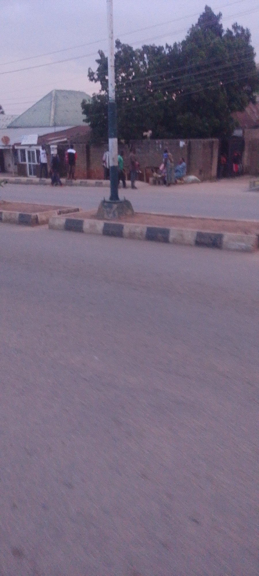 The image depicts a street scene in Gura Top, Nigeria. A light pole with blue and white stripes stands in the foreground. Beyond the pole, a group of people are gathered near some buildings. There are several structures visible, including a building with a green, triangular roof and another with a brown facade. Some individuals appear to be engaged in commerce, with goods displayed on the ground. The road surface is paved, and a curb with black and white markings separates it from the sidewalk area where the people are. Trees with dense foliage are present in the background, and power lines are strung overhead. The sky is overcast and a muted purple hue, suggesting it is either early morning or late evening.