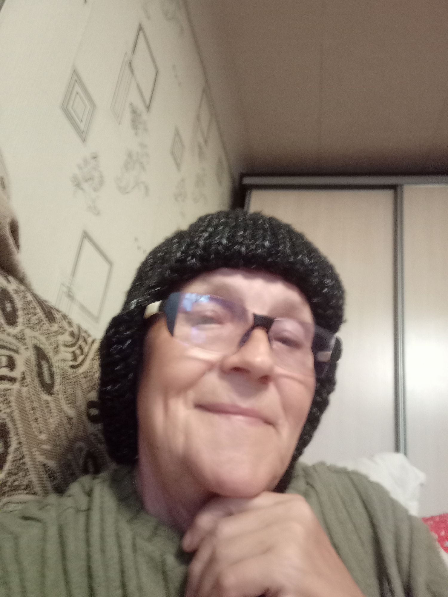 A woman is taking a selfie. She is wearing a black knitted hat and glasses. She is smiling and resting her chin on her hand. The background appears to be a room with patterned wallpaper and a closet. There is no information available about the location in the city of Degtyarsk, Russia.